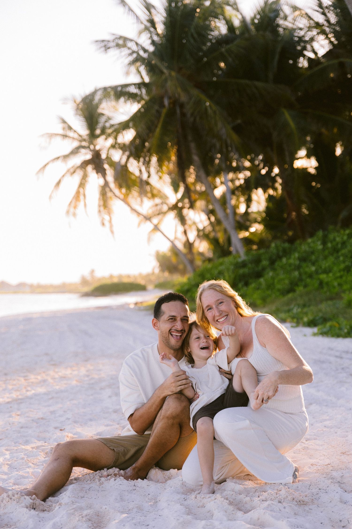 Happy family of three laughing together on a tropical white sand beach at sunset with palm trees in the background.