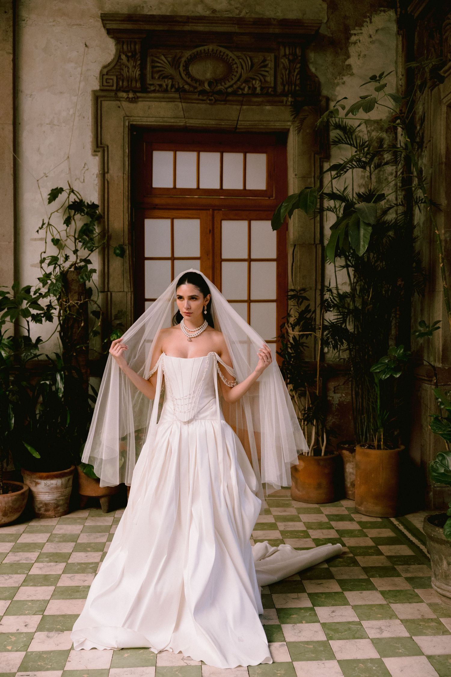 Person in a flowing white wedding dress posing in a tiled courtyard with potted plants.