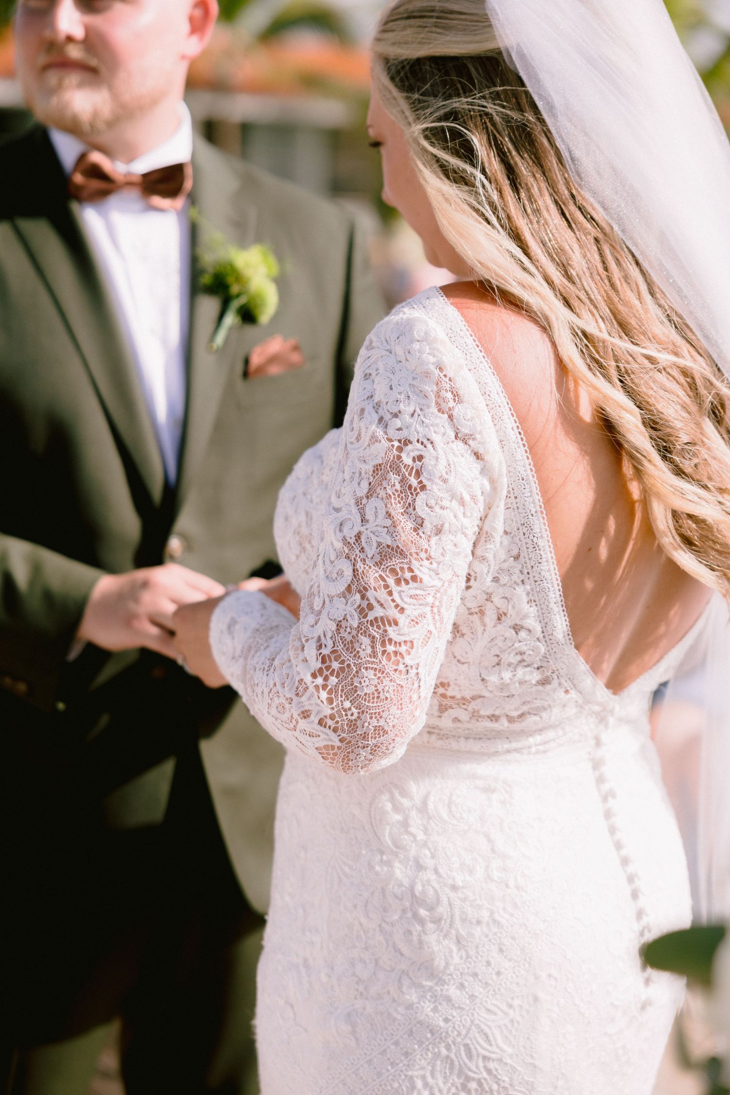 Bride in lace long-sleeve wedding dress with open back exchanges rings with groom in olive suit and bow tie.