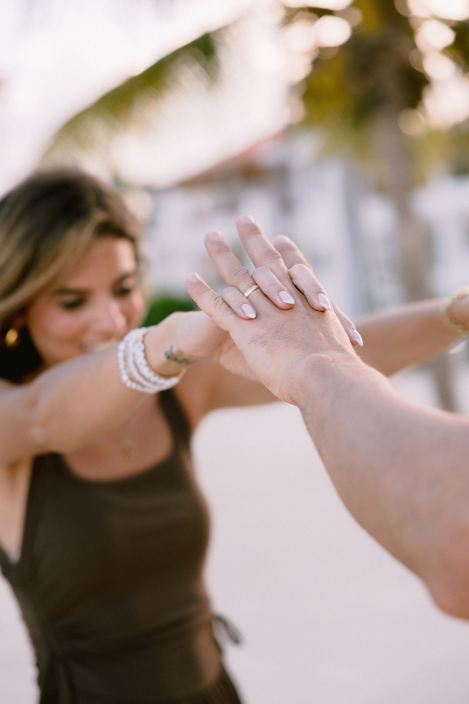 Hands displaying engagement rings and wedding bands in a soft focus outdoor setting.