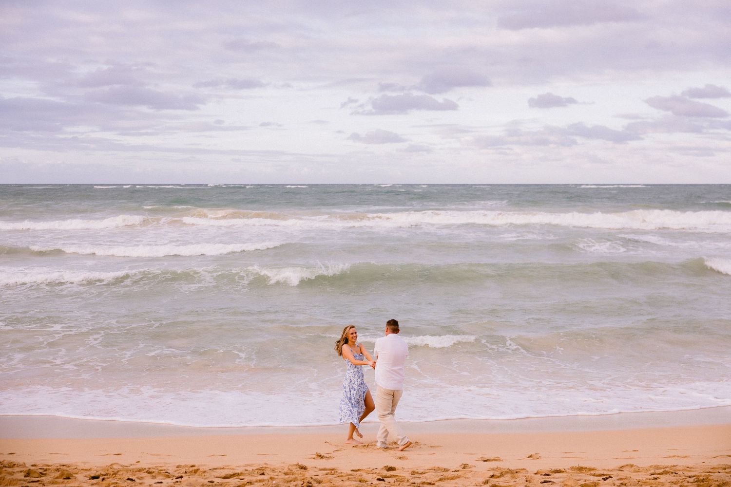 A couple walks hand in hand along a sandy beach with rolling ocean waves under a cloudy sky.