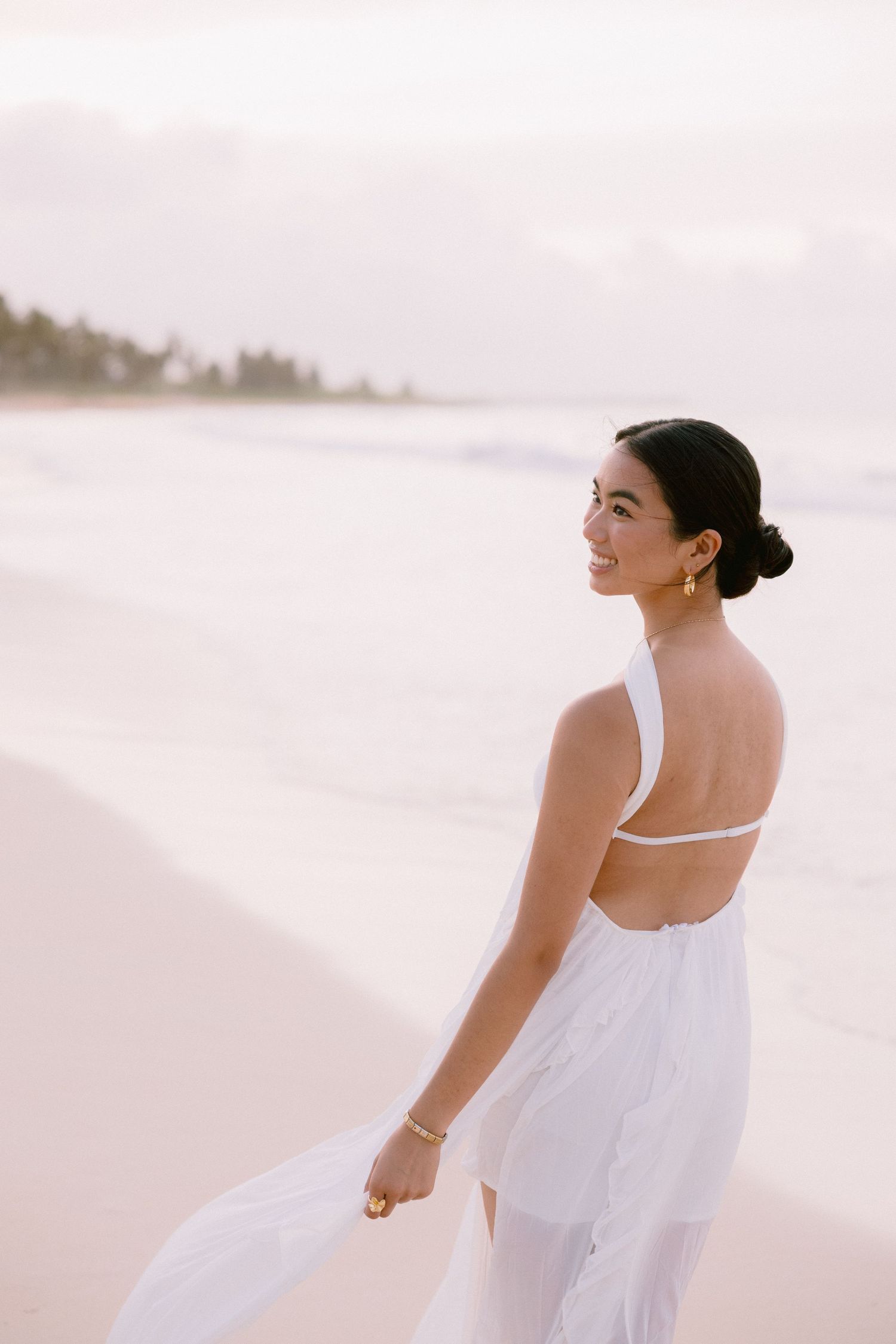 A smiling woman in a white backless one-shoulder dress stands on a sandy beach at sunset.