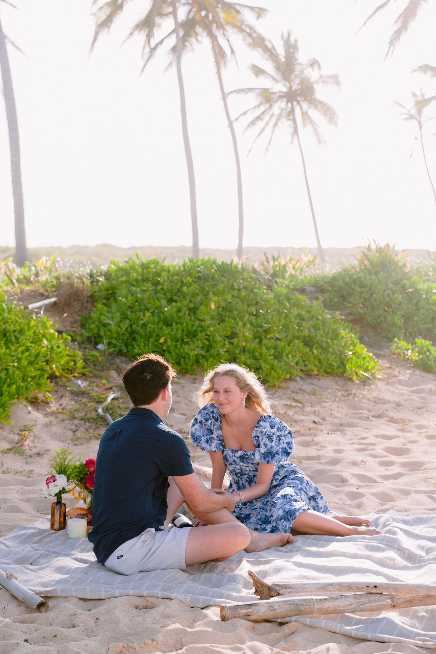 Couple lying on a white blanket on a sandy beach with palm trees in the background.