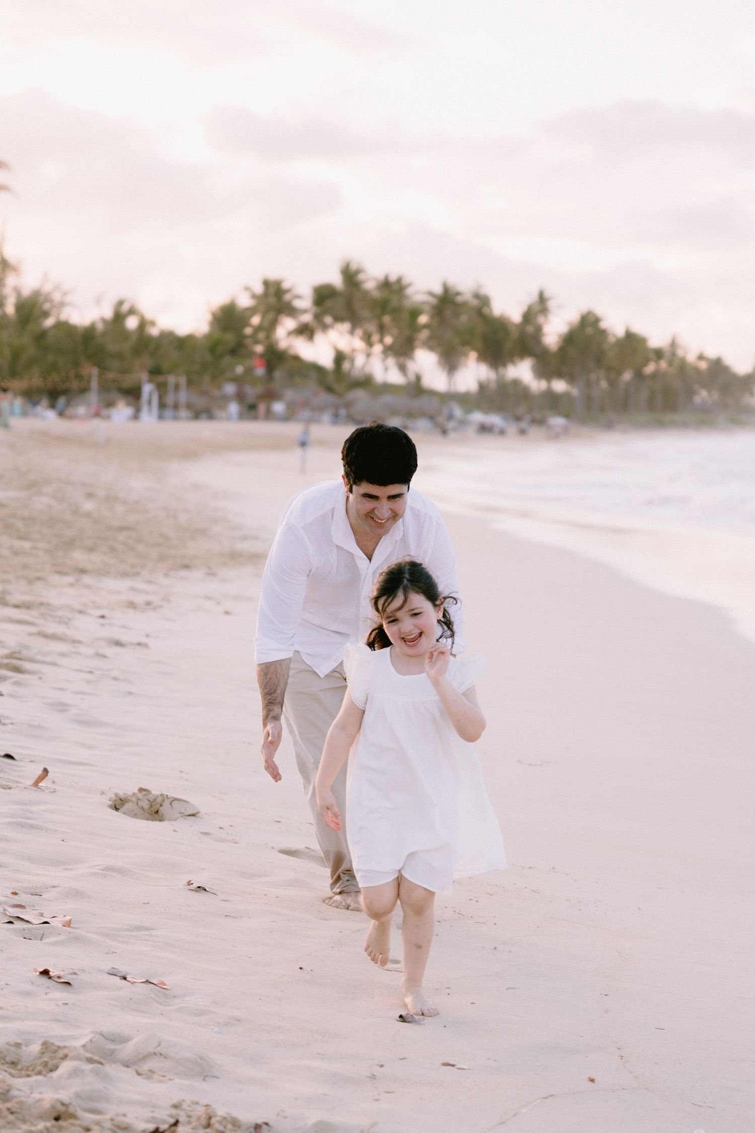 A couple in white clothing playfully runs and embraces along a serene tropical beach at sunset.