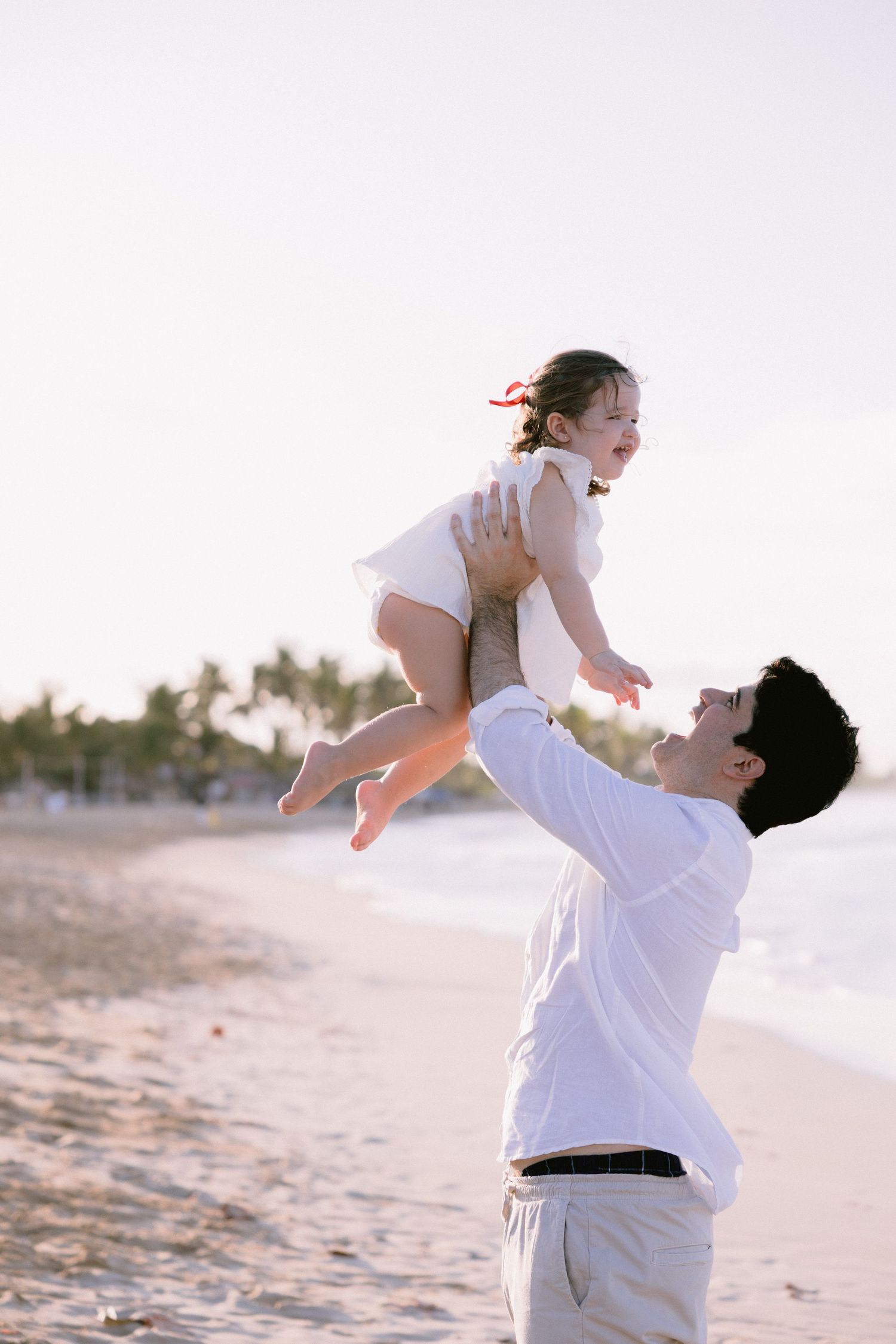 A joyful moment captured on a beach as a parent lifts a child in a white dress during sunset.