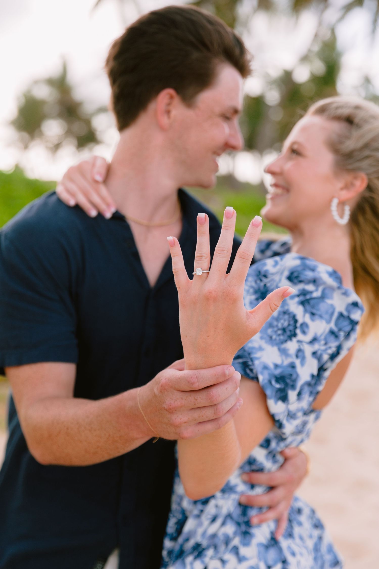 A couple in blue and navy outfits show off their engagement ring in a series of romantic beach photos.