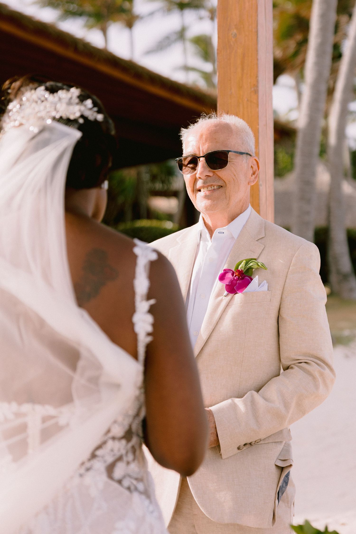 A couple in light-colored wedding attire smiles during an outdoor ceremony with palm trees in the background.
