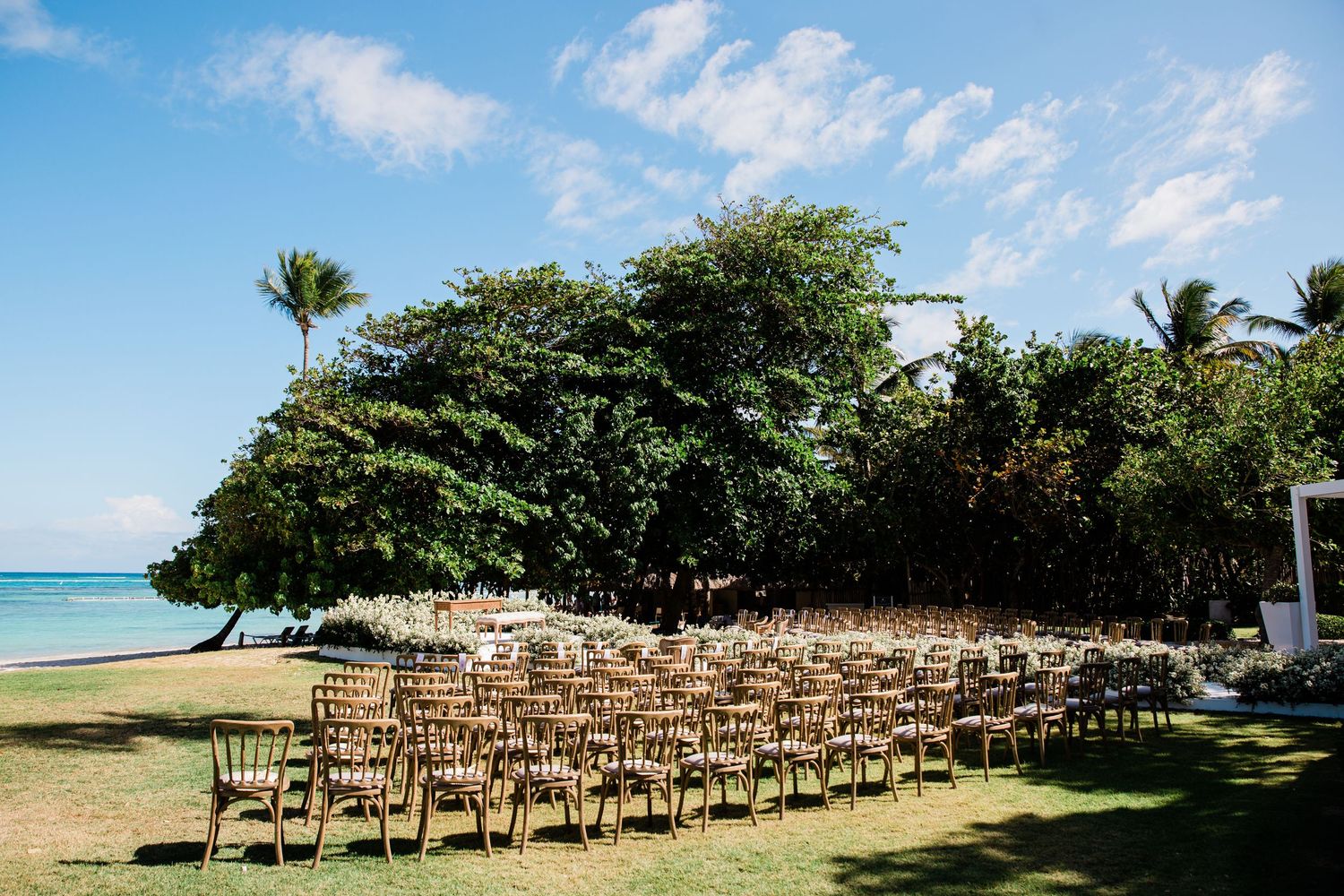 Wooden folding chairs arranged in rows on a lush green lawn overlooking a tropical beach for an outdoor wedding ceremony.