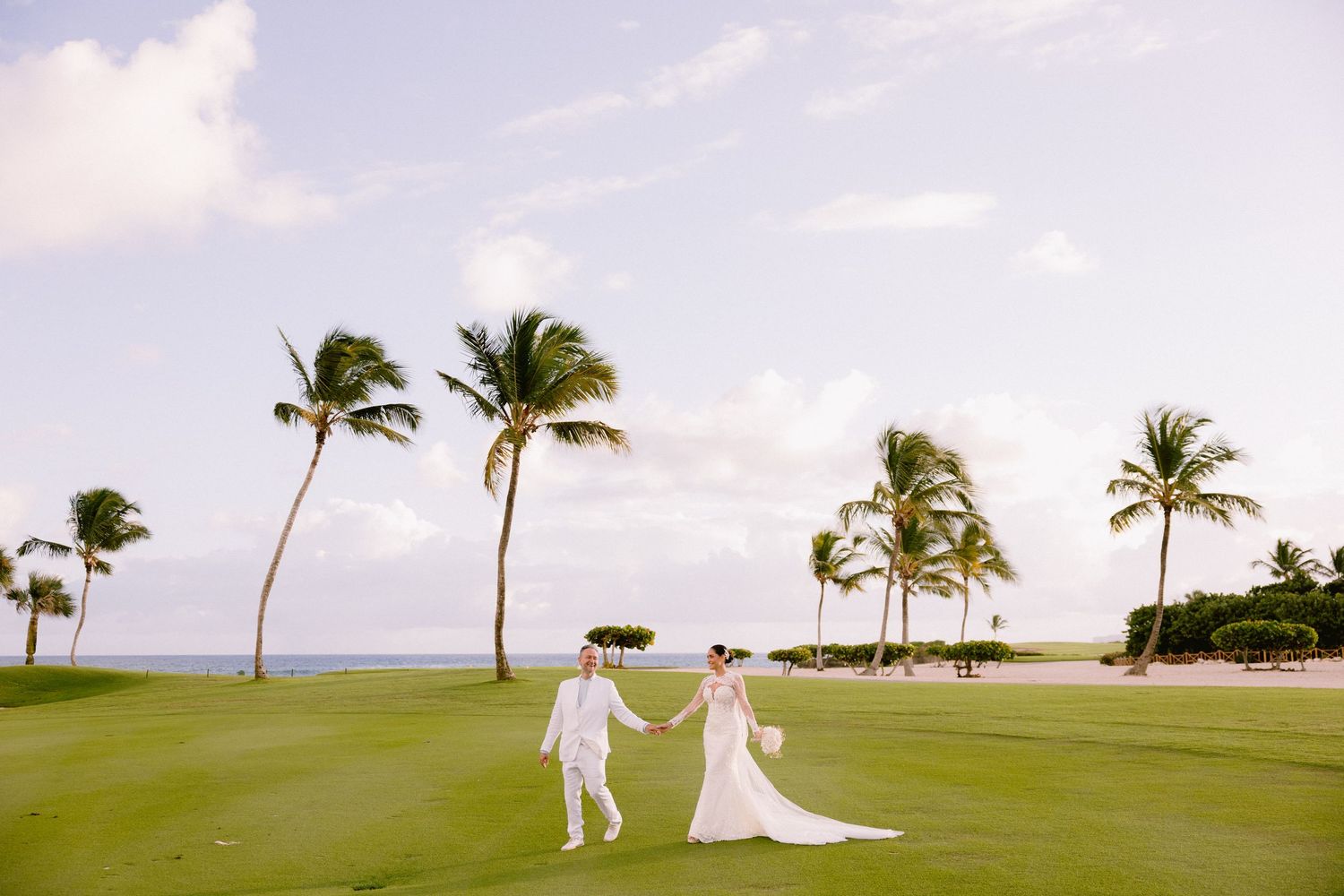 Bride and groom walk hand-in-hand on lush green golf course with palm trees and ocean view on a sunny day.