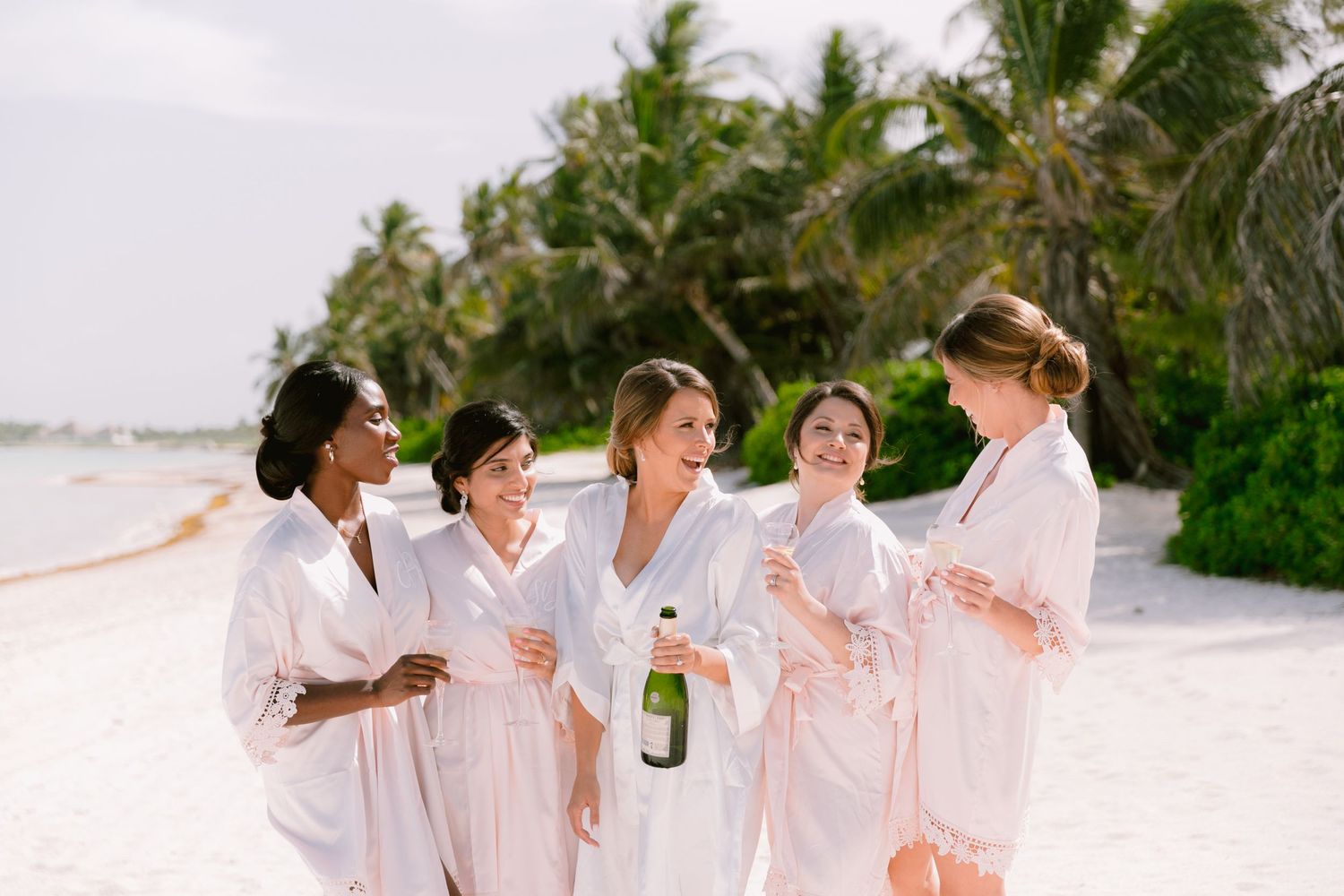 Five bridesmaids in white robes celebrate on a tropical beach with champagne, surrounded by palm trees.