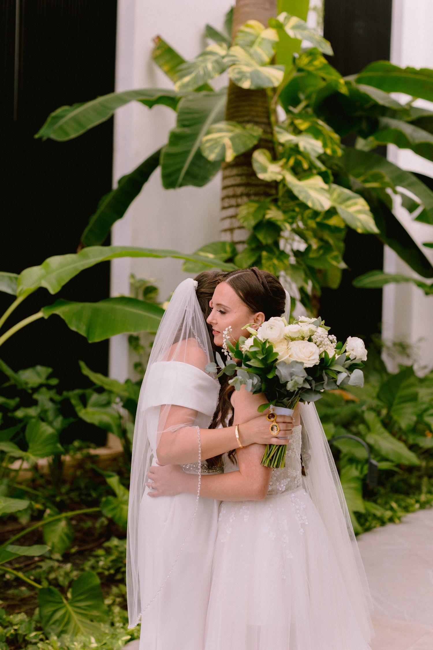 Bride in white off-shoulder gown and veil holds white floral bouquet, embracing someone amid lush tropical greenery.