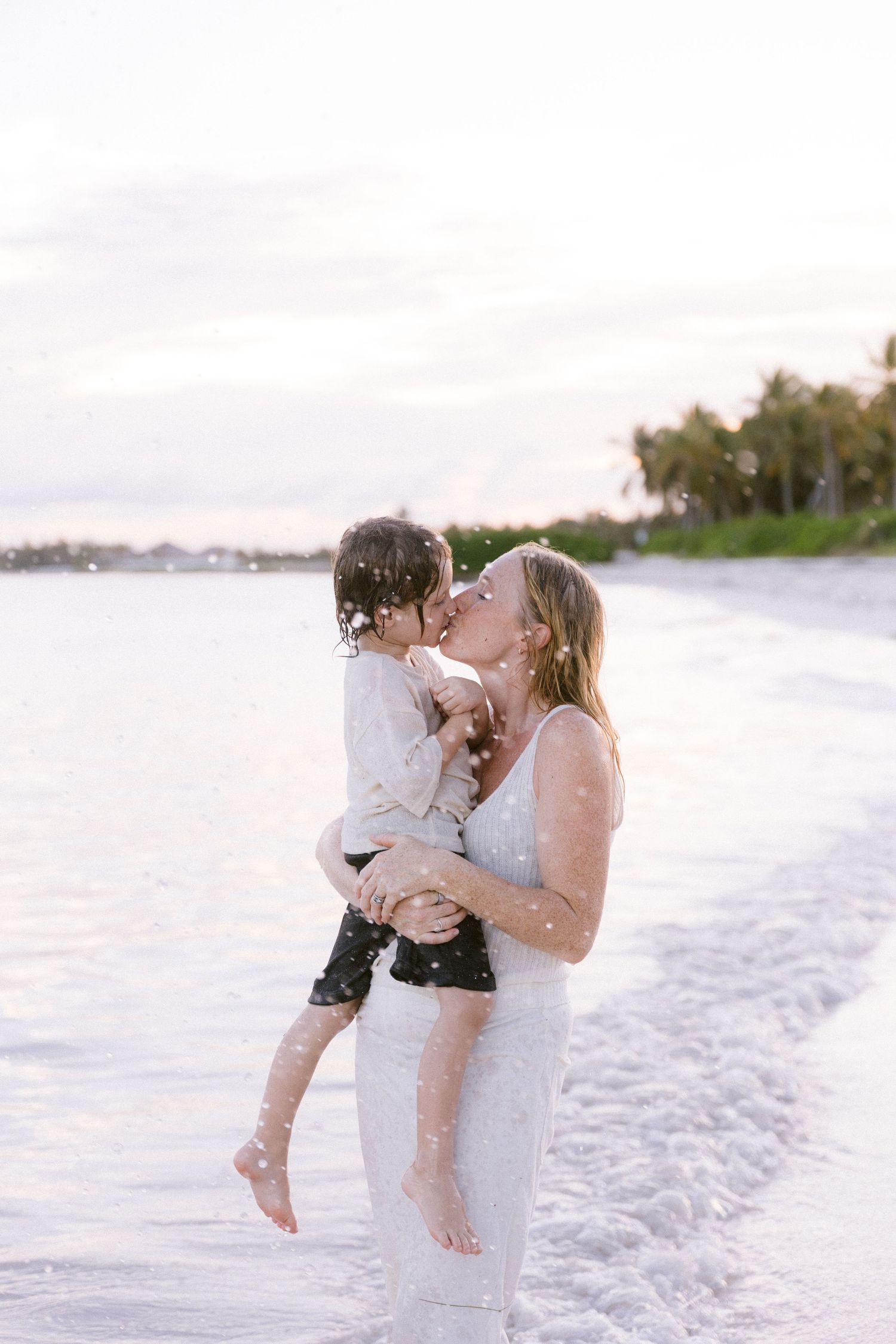 A mother kisses her young child while standing in ocean waves on a tropical beach at sunset.