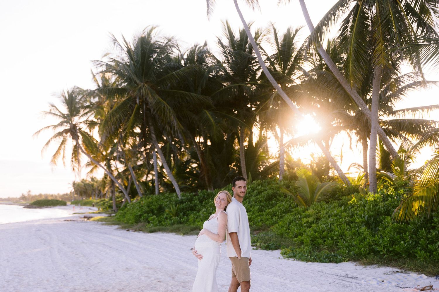 Couple in white attire walking on a tropical beach at sunset with lush palm trees in the background.