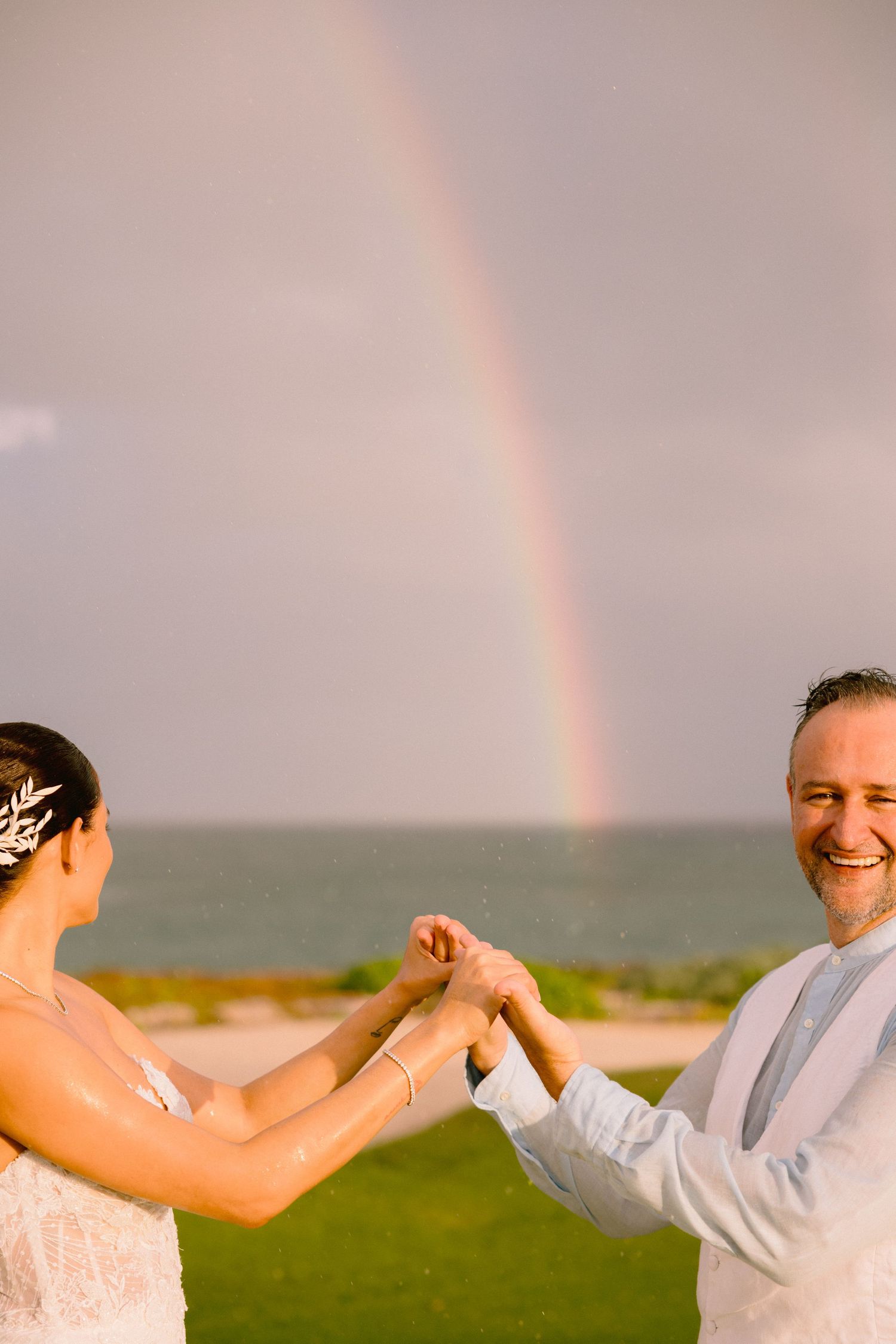 A couple dances together on a grassy oceanside cliff with a vibrant rainbow arching across the cloudy sky in the background.
