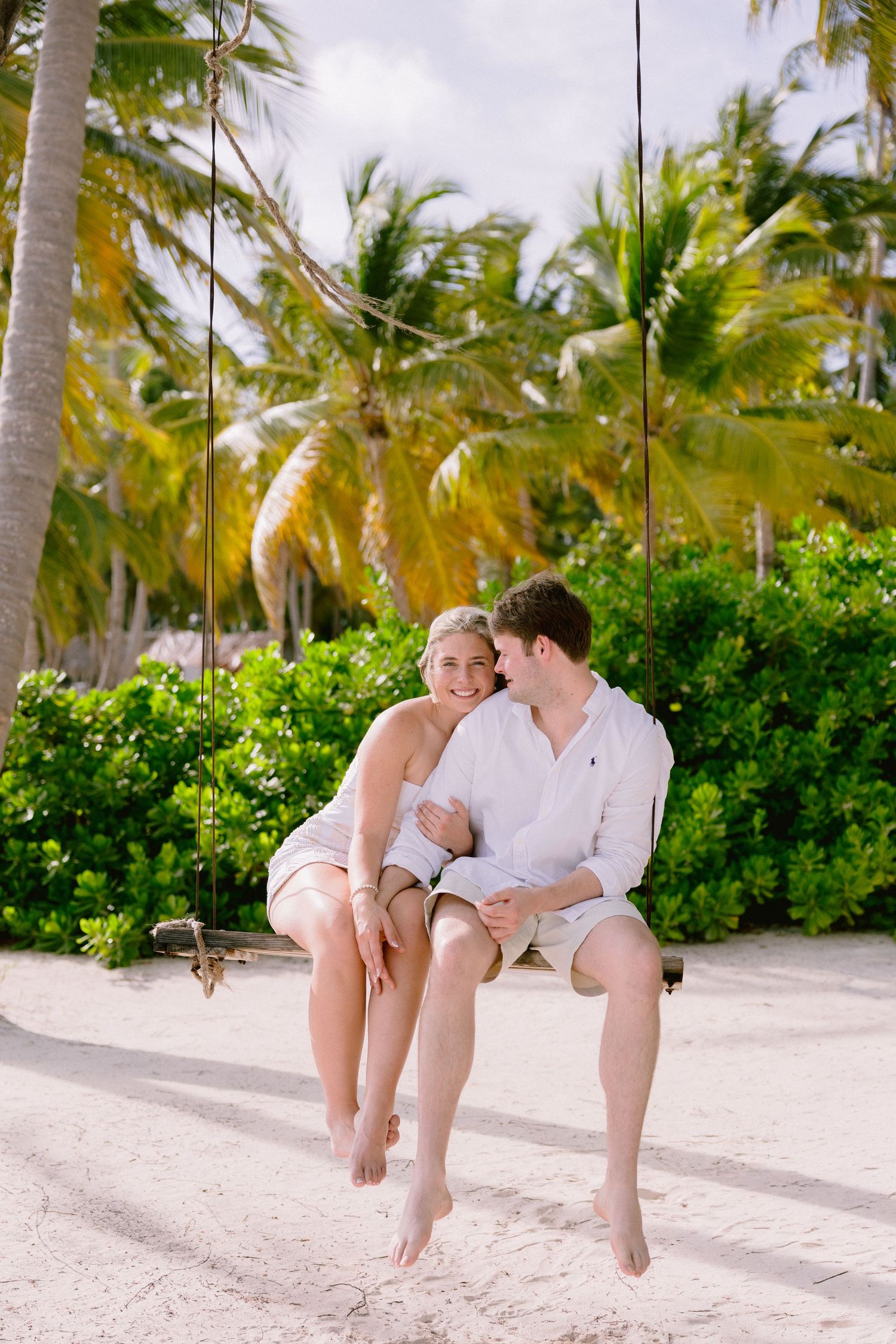 A couple sits together on a white sandy beach with palm trees and tropical foliage in the background.