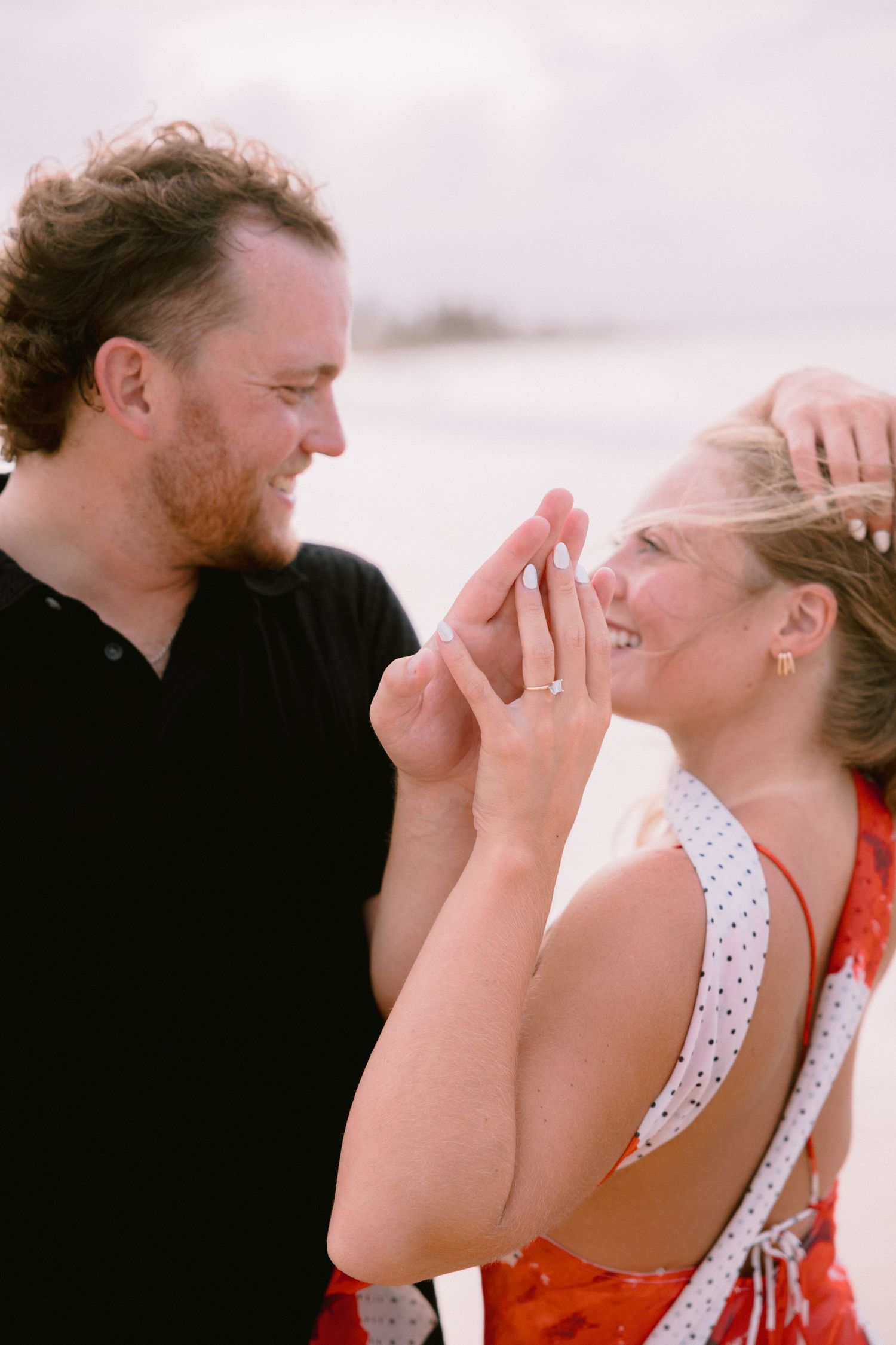 Couple smiling at each other on a beach, woman showing off engagement ring in a red dress.