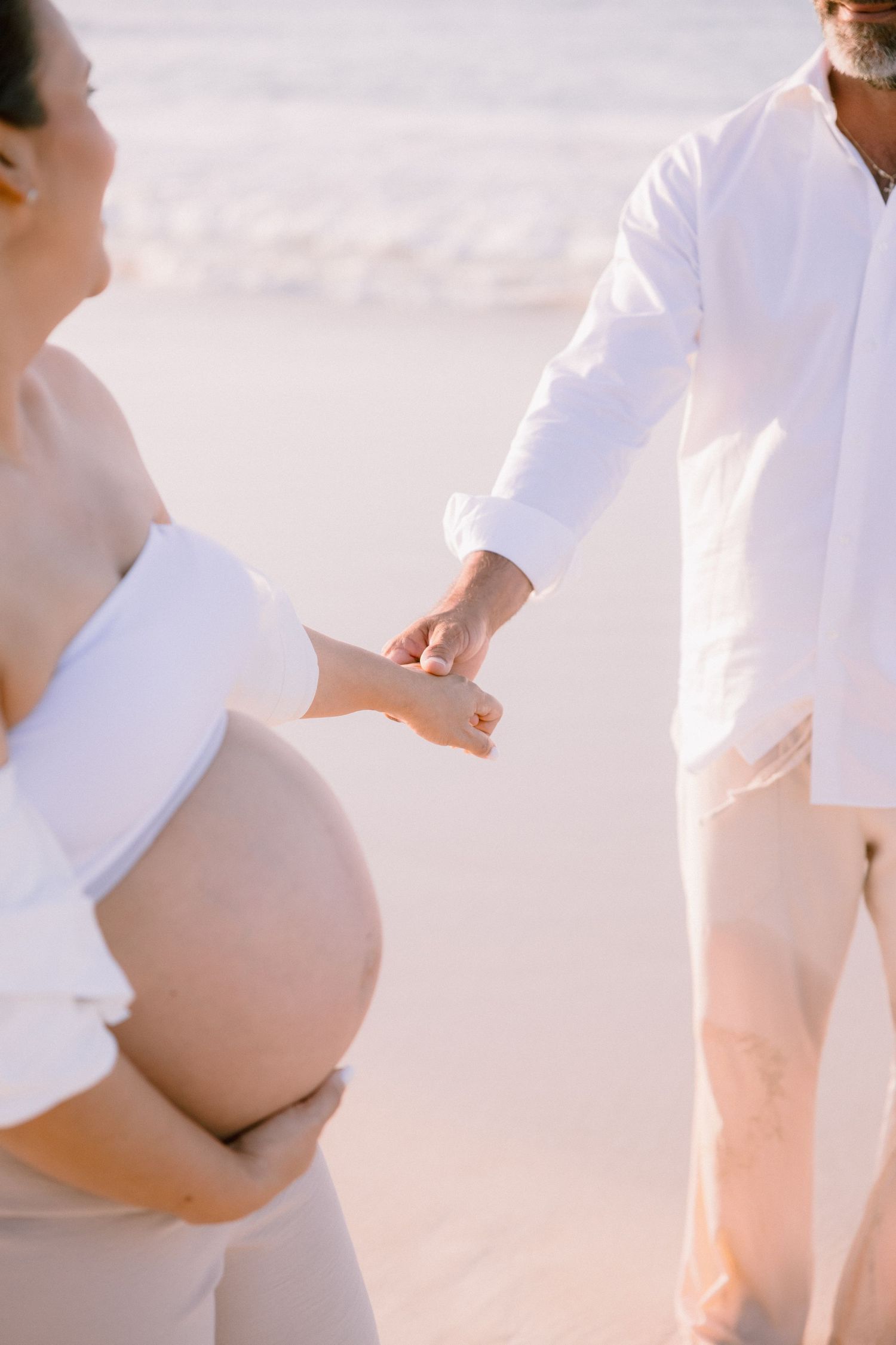 Pregnant couple holding hands on a beach, woman in white crop top cradling baby bump, man in white shirt, soft ocean backdrop.