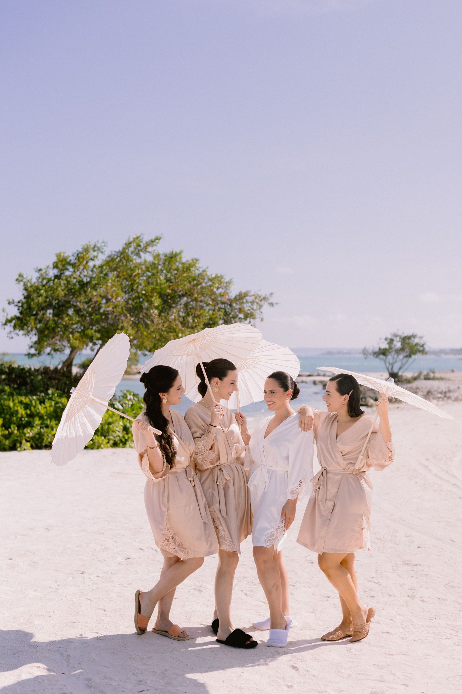 Group wearing matching beige outfits poses playfully on a white sandy beach with ocean and trees in background.