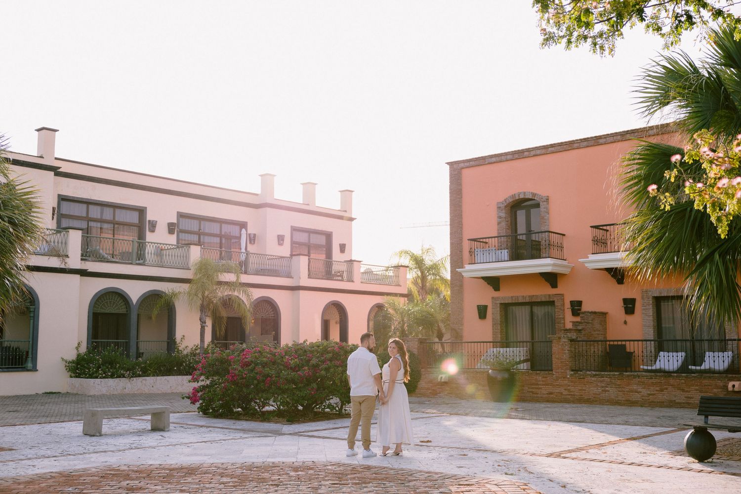 Couple standing in a sunlit courtyard surrounded by colorful colonial-style buildings and palm trees.