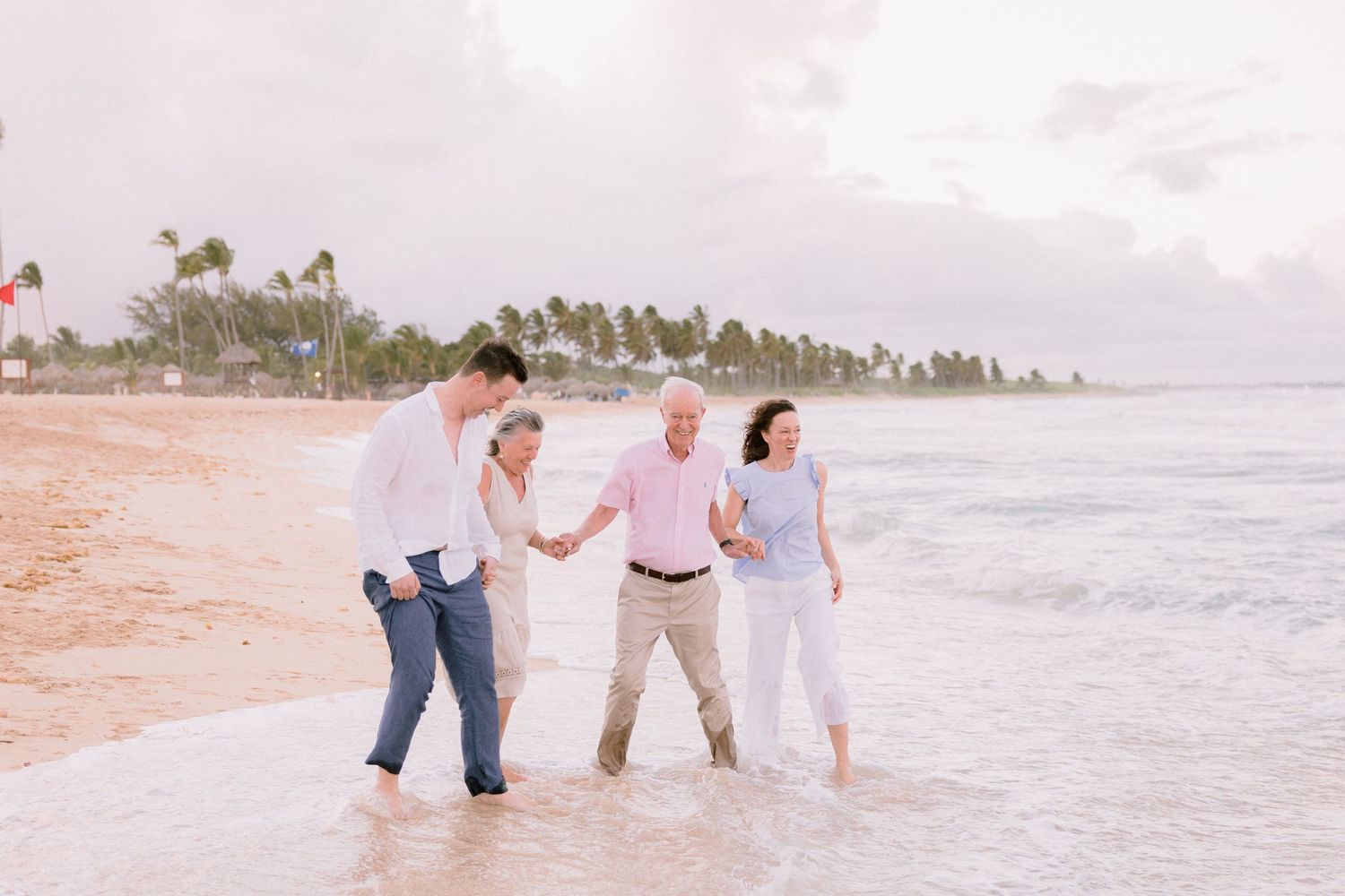 Two couples walk hand-in-hand along a tropical beach shore, waves washing over their feet at sunset.