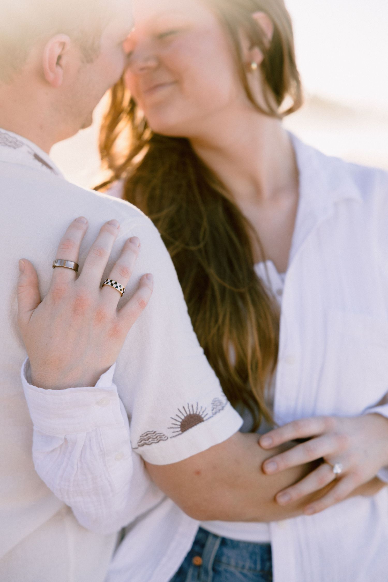Couple embracing closely, woman wearing engagement ring and wedding band, both in white shirts, romantic outdoor photo.
