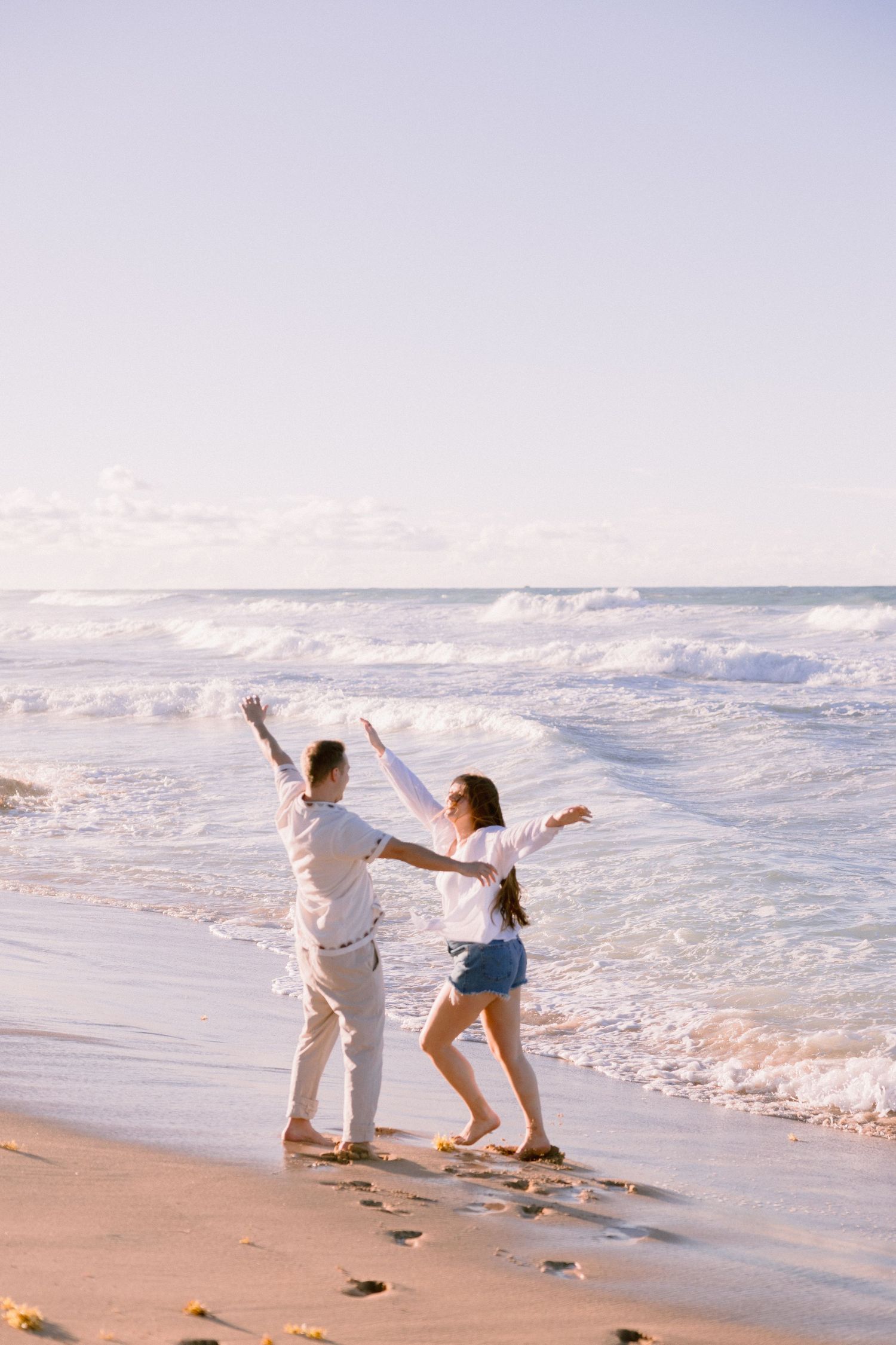 Couple dancing joyfully on a sandy beach at sunset with ocean waves crashing behind them.