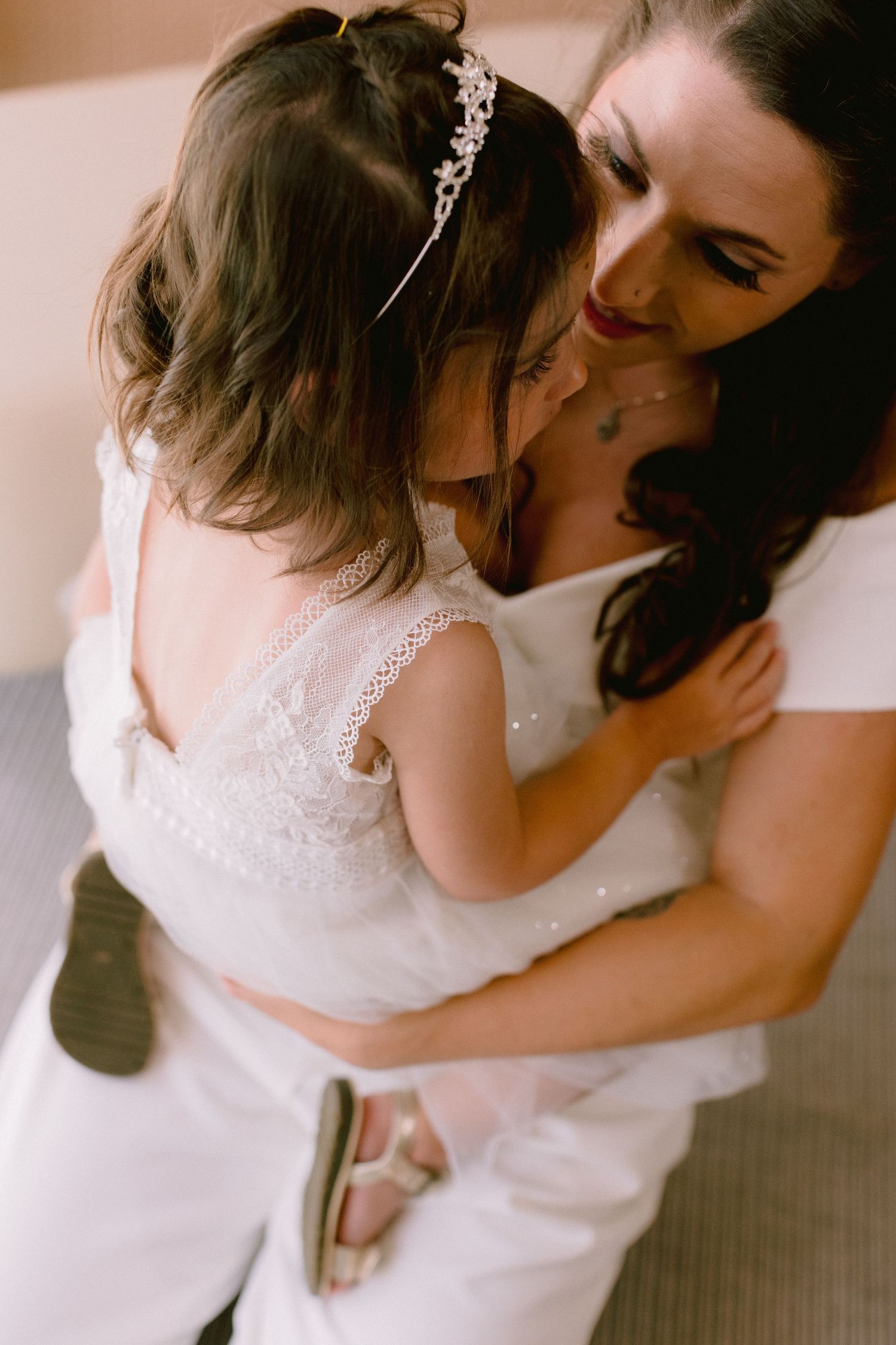A bride in white holds a young flower girl in a lace dress with a crystal headband, sharing a tender moment.