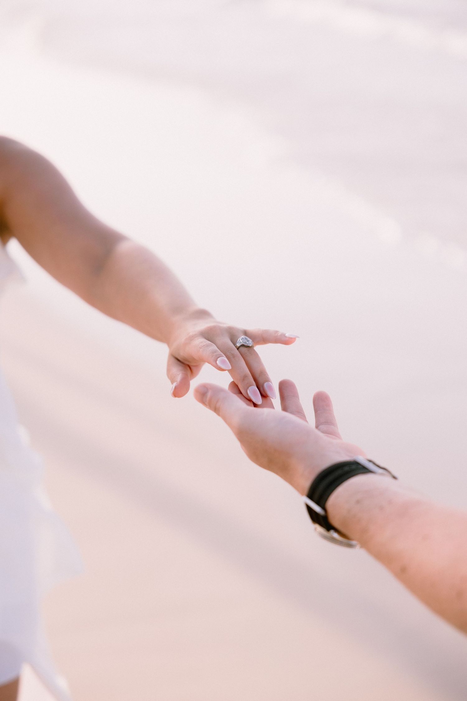 Two hands reaching toward each other on a beach, woman wearing a diamond engagement ring, man with black watch.