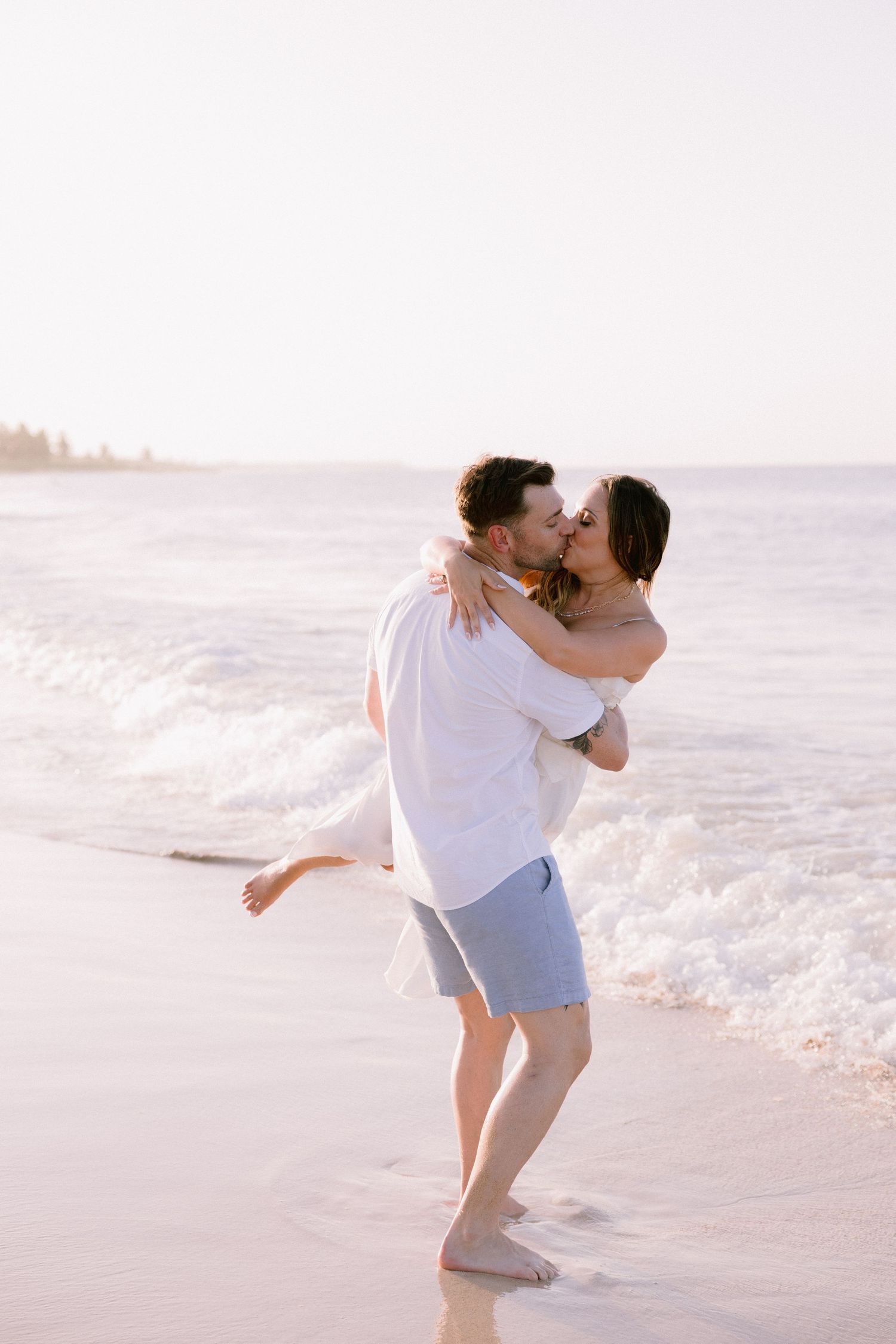 Couple kissing on a beach at sunset, man lifting woman in white dress near ocean waves.