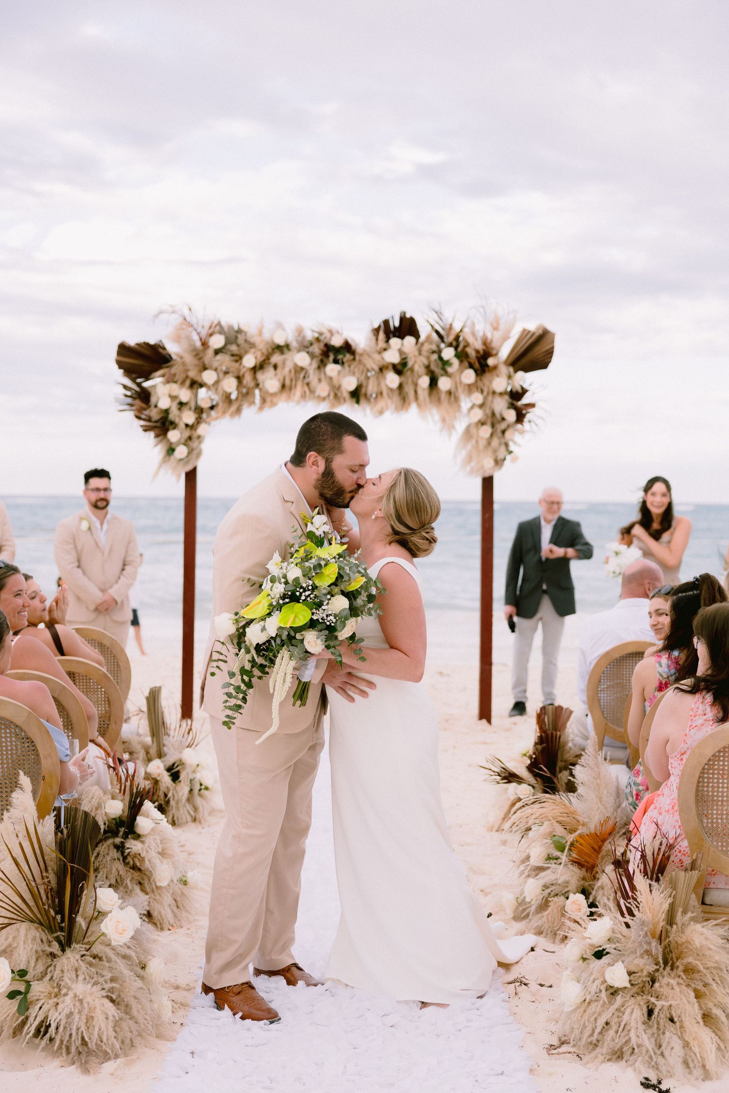 Couple sharing first kiss at beach wedding ceremony under floral arch with guests seated on wicker chairs.