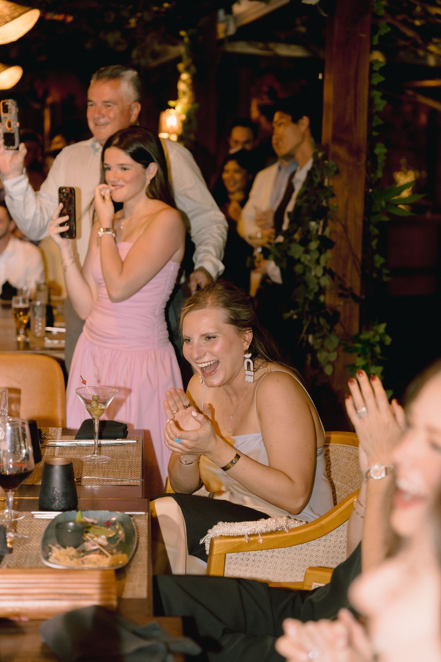 Guests laugh and clap at a lively dinner reception while a woman in a pink dress films on her phone beside a cocktail-topped table.