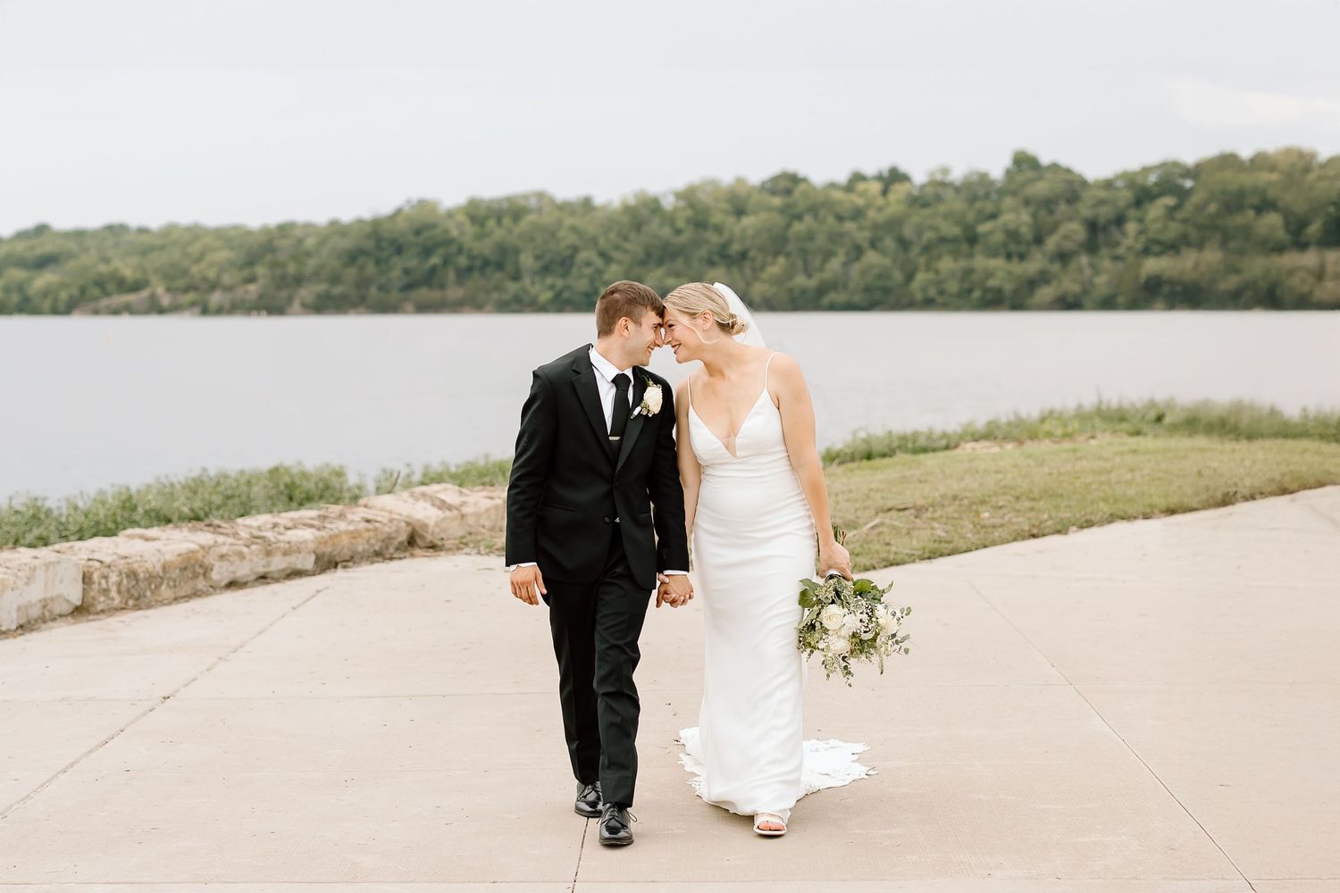 Couple walking together on a winding dirt path near a lake surrounded by green hills and trees.
