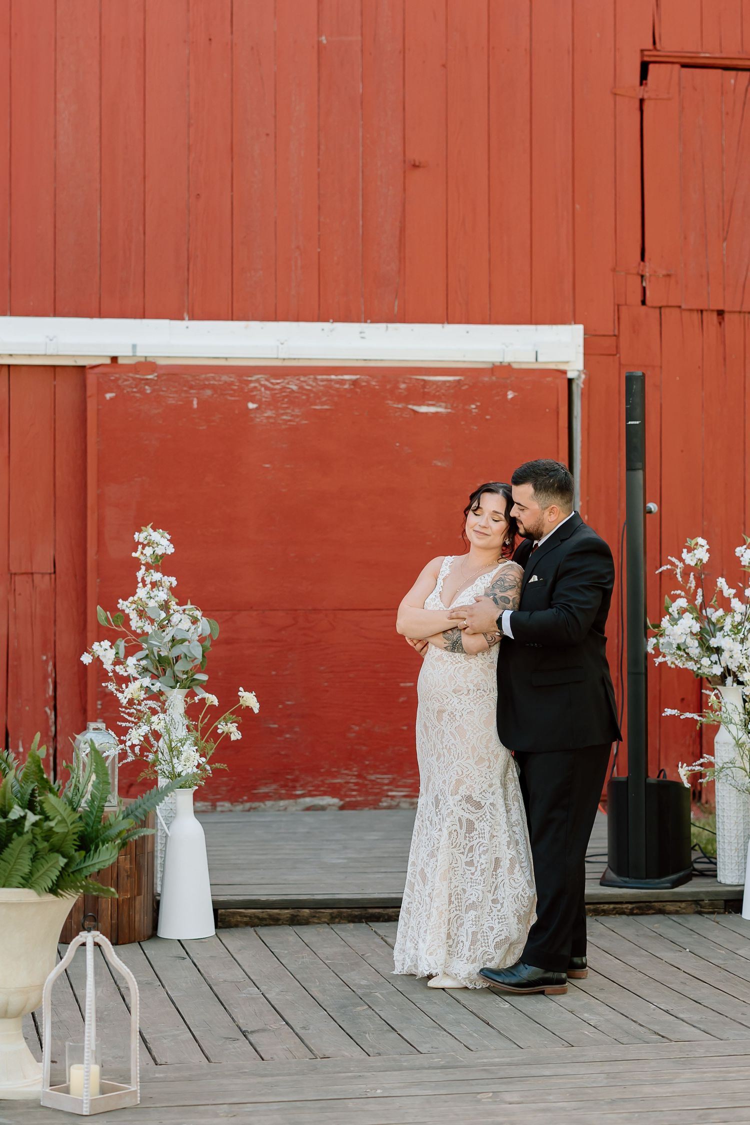A romantic wedding couple shares tender moments against a rustic red barn wall decorated with white floral arrangements.