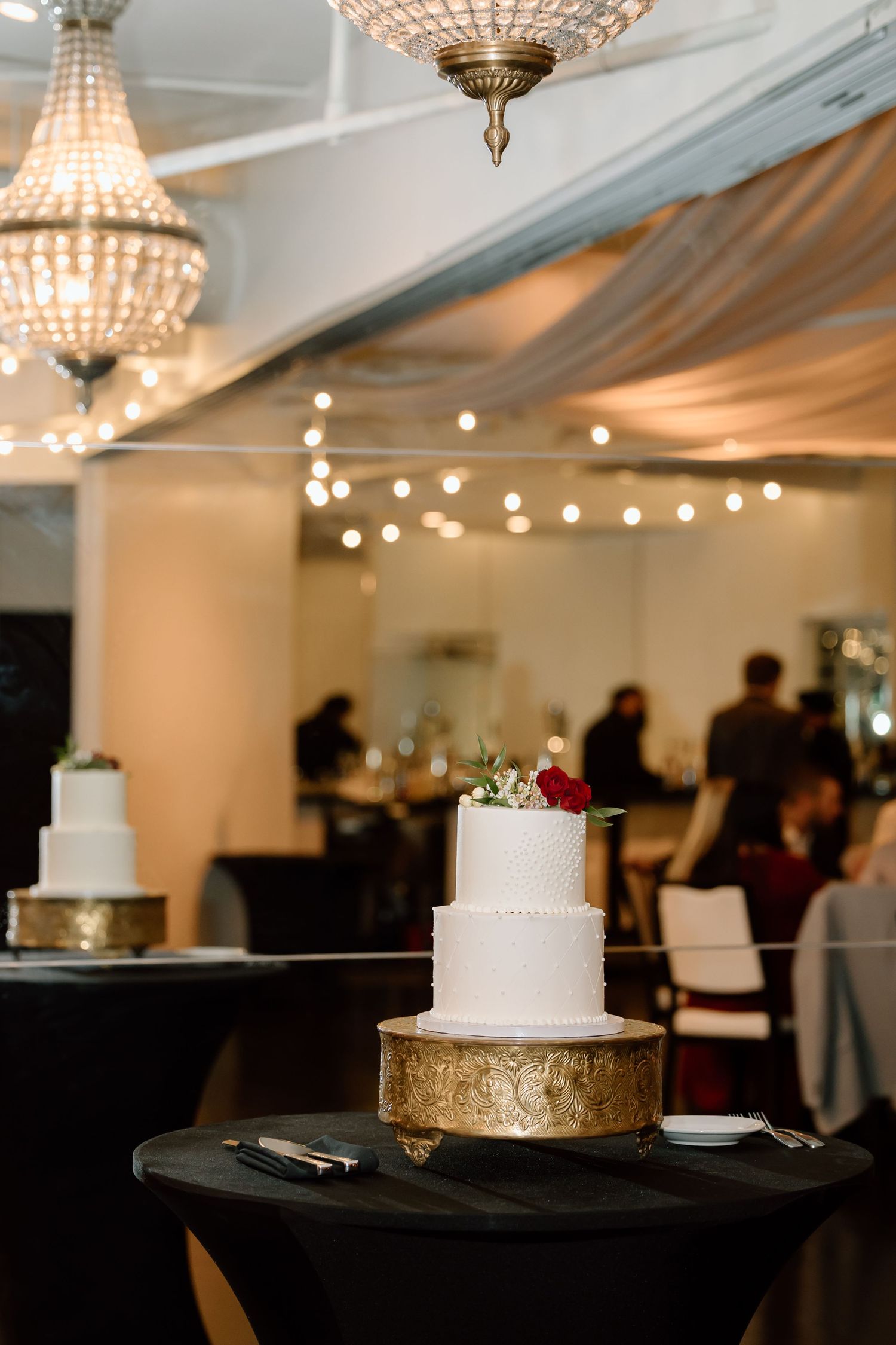 Elegant white wedding cake displayed on gold stand under crystal chandeliers at an indoor reception venue.