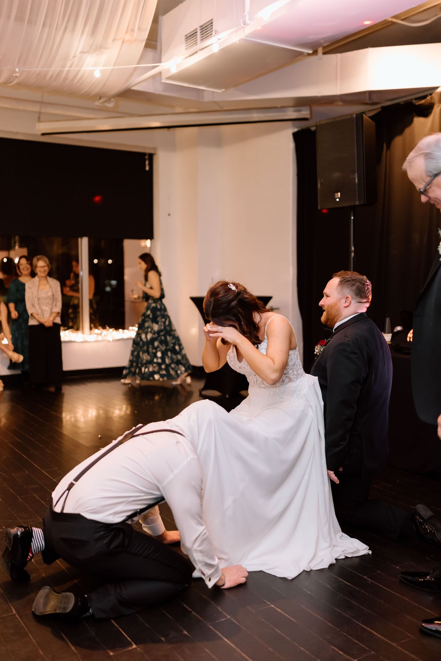 Wedding party members participate in a fun limbo dance game at an indoor reception venue with dark wood floors.
