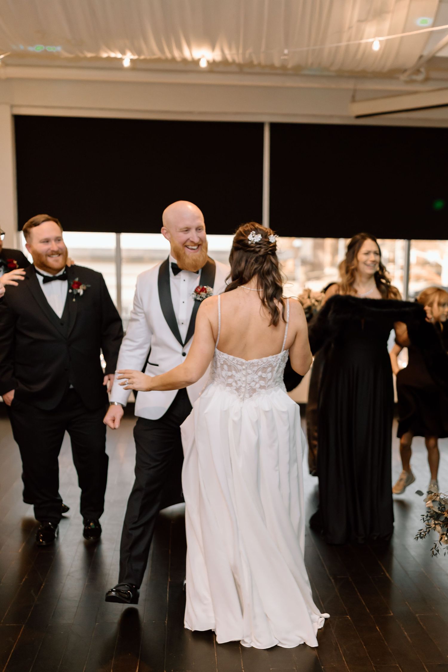 Wedding reception guests dance on a dark hardwood floor under warm lighting at an indoor venue.