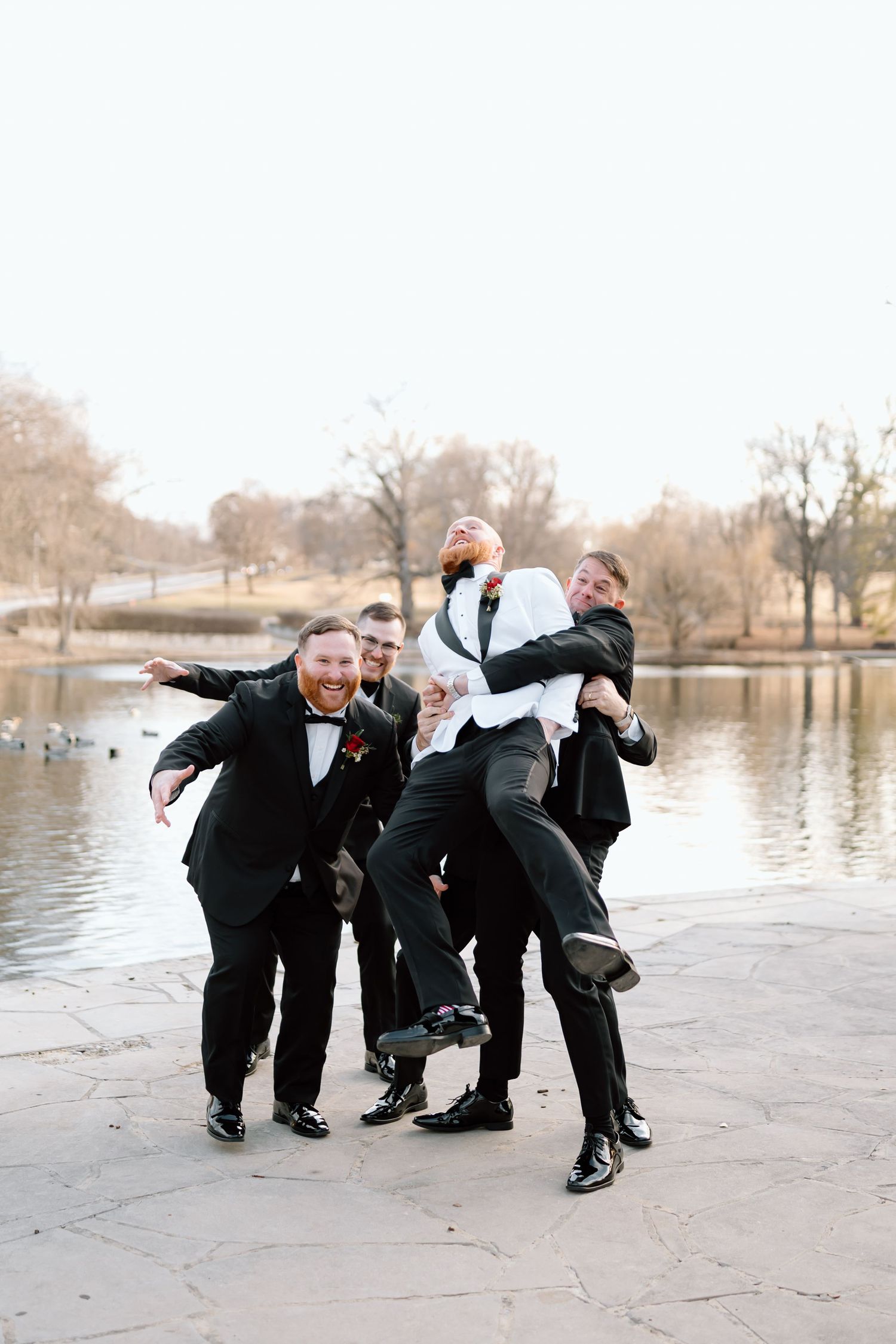 Wedding party members in black tuxedos pose playfully by a lake on a winter day.