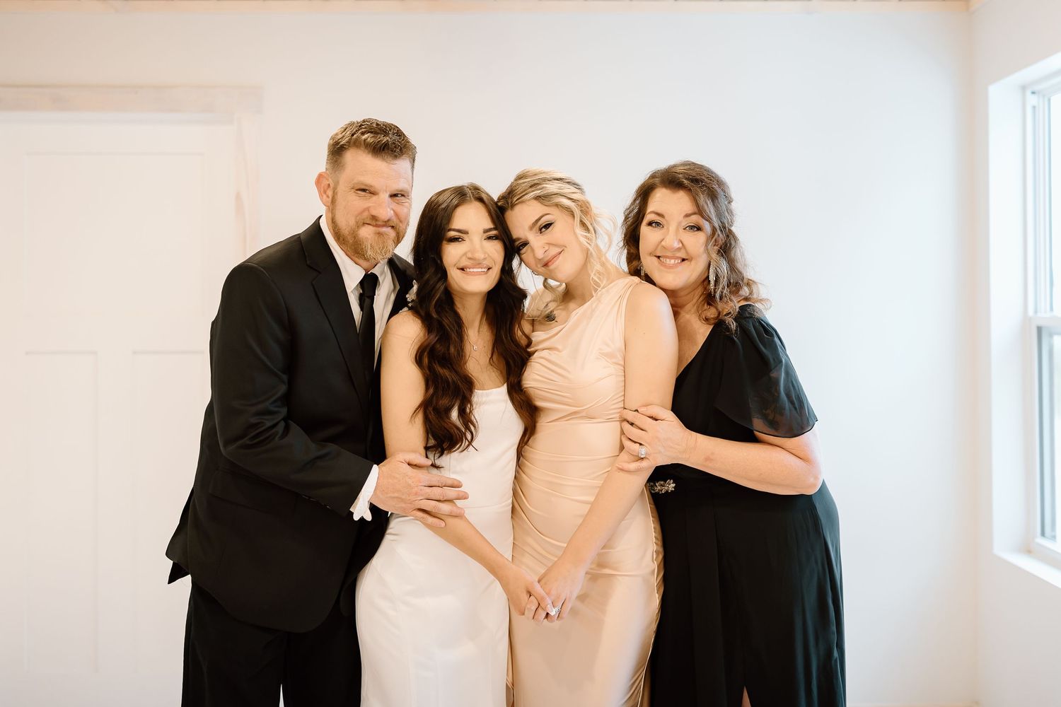 A joyful group embraces and laughs together while wearing formal attire in a bright white room.