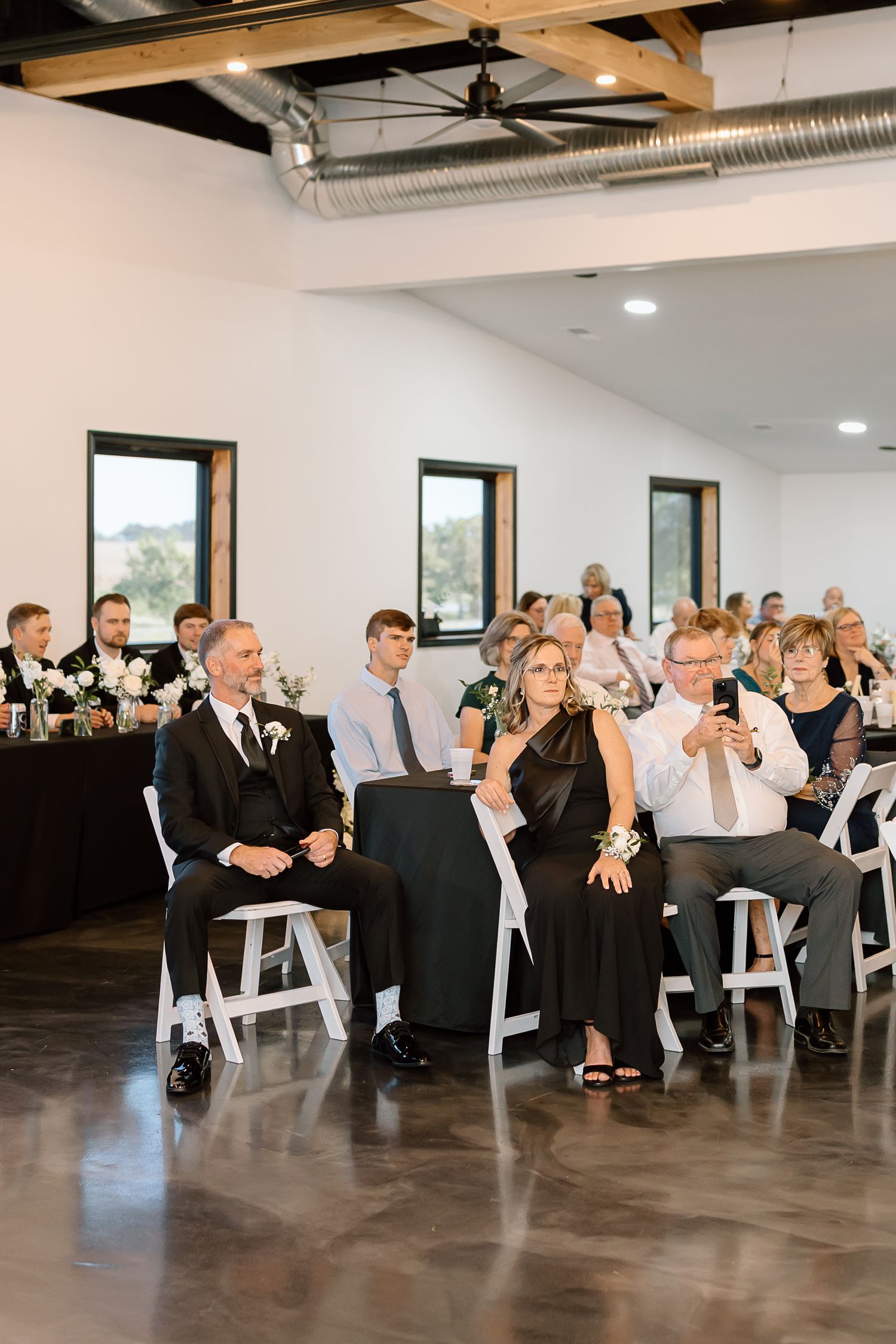Wedding guests seated in white chairs watch an event in a modern venue with exposed ductwork and large windows.