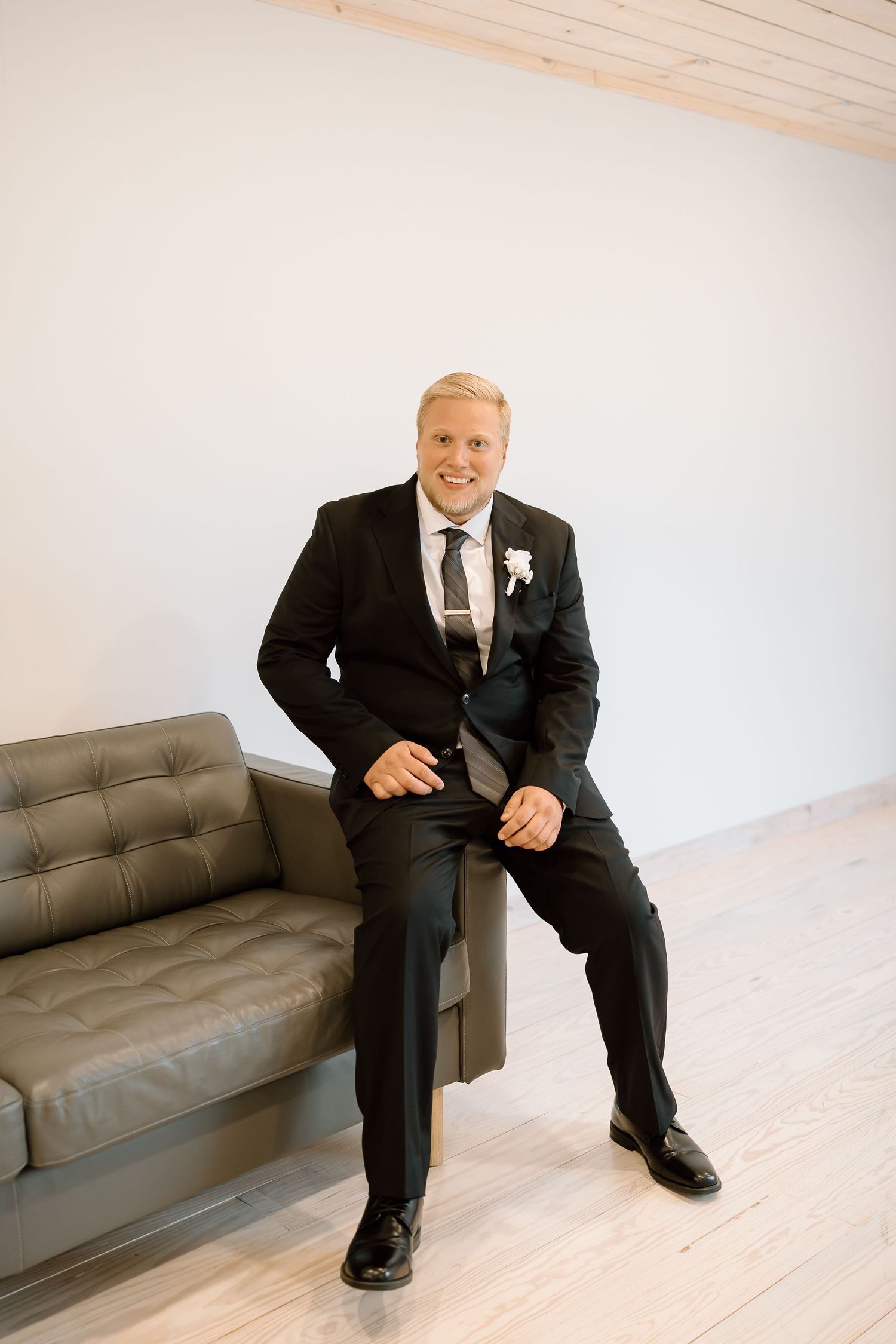 A groom in a classic black suit and tie sits on a gray leather couch in a bright minimalist room.