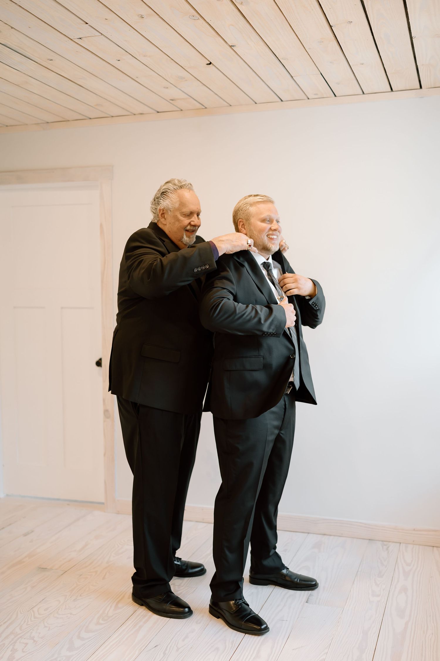 Two figures in black suits stand in a minimalist white room with wooden ceiling panels.