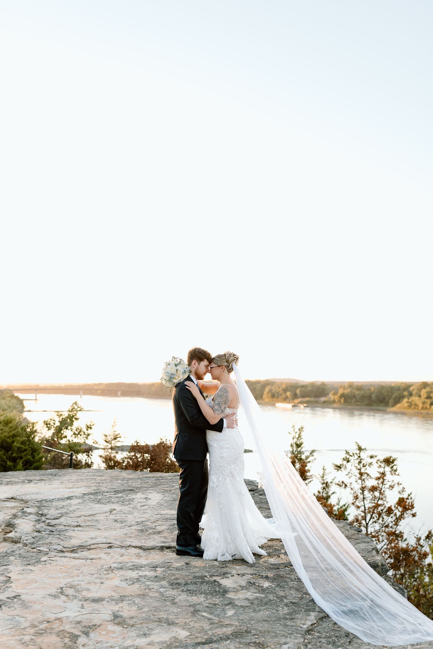 Newlywed couple embraces on rocky cliff overlooking scenic lake at sunset with flowing white veil in the breeze.