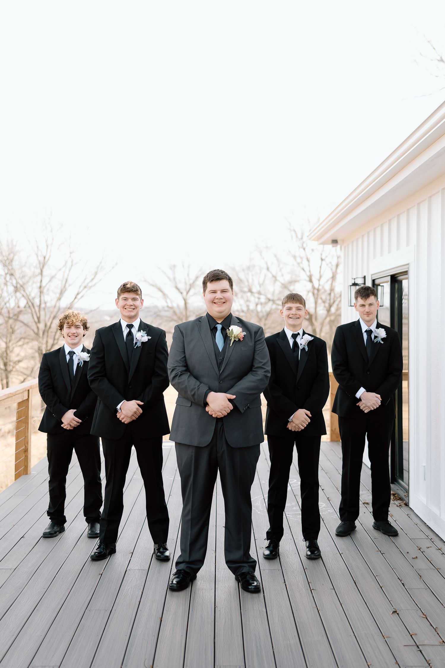 Groomsmen in black suits and ties pose together on a white wooden deck for a formal wedding photo.