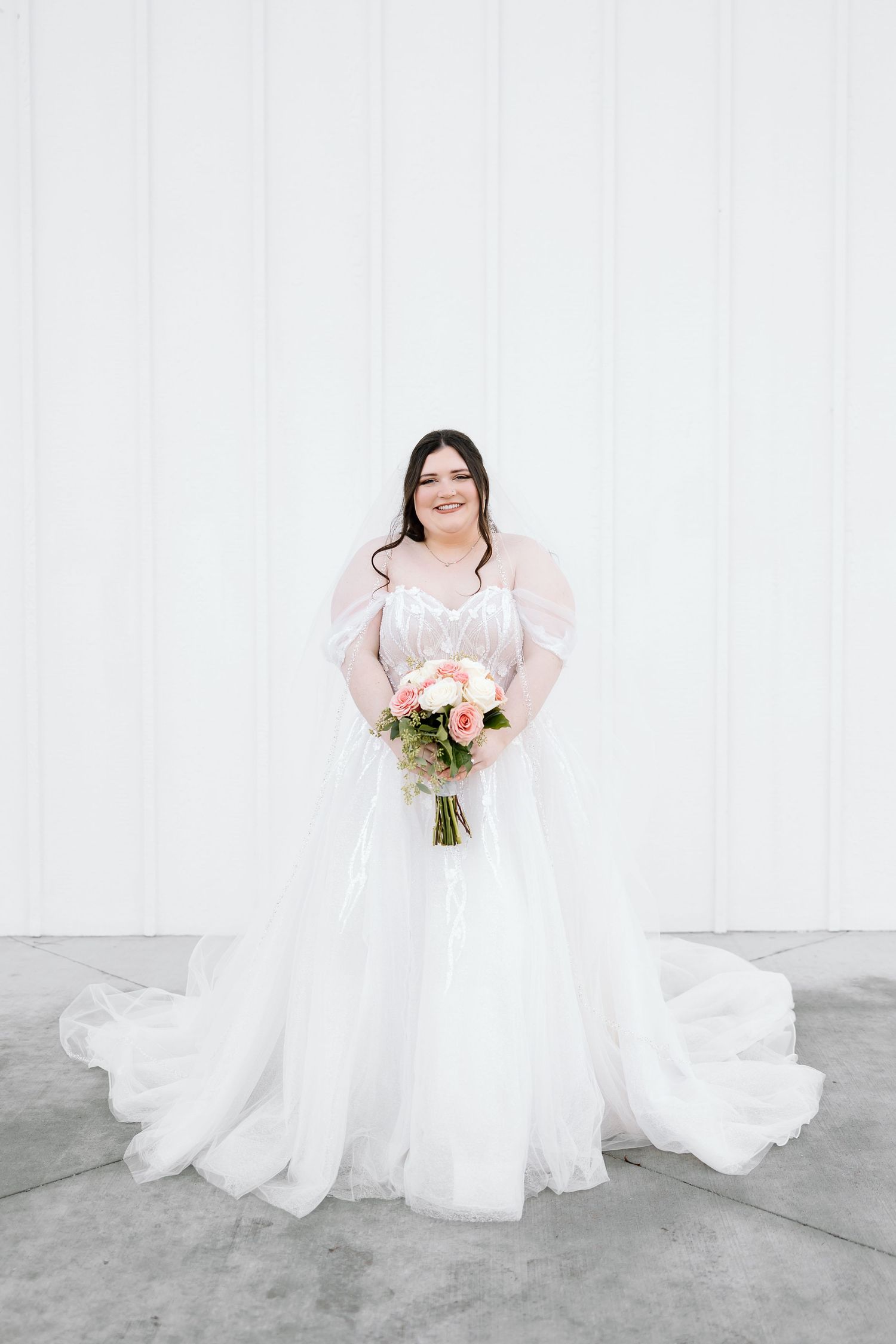 A bride in a flowing white wedding gown holds a pink and white bouquet against a light gray backdrop.