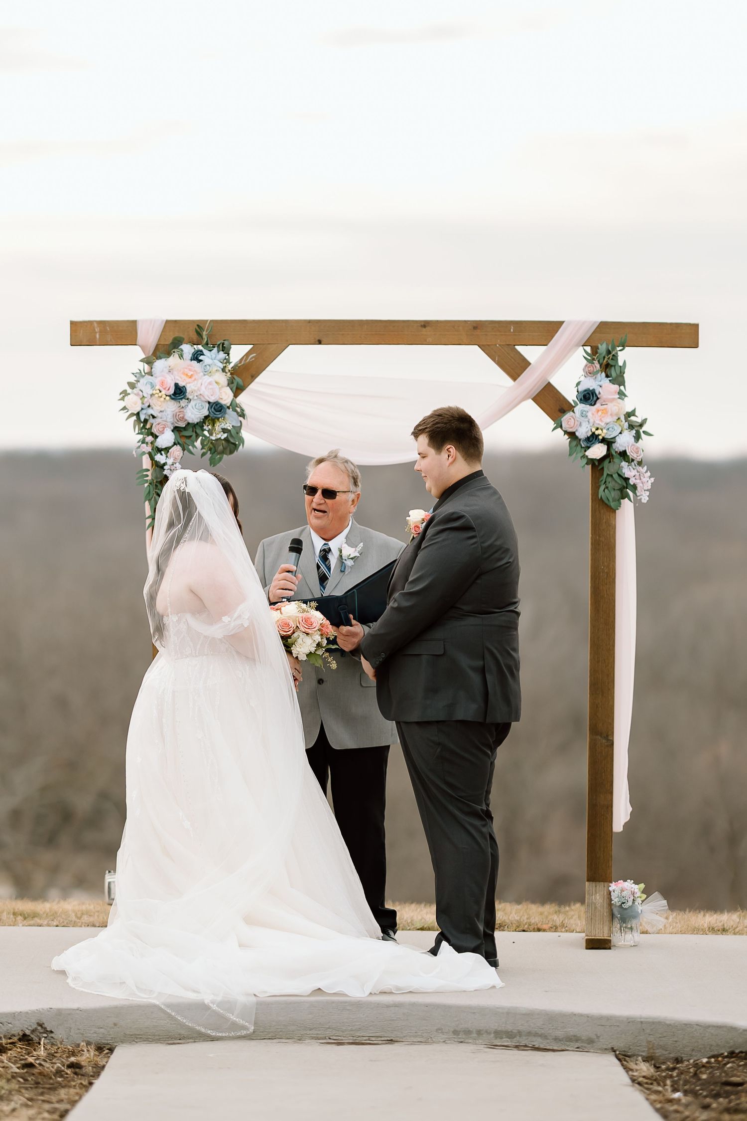 A wedding ceremony takes place at an outdoor venue with a wooden arch decorated with white floral arrangements.