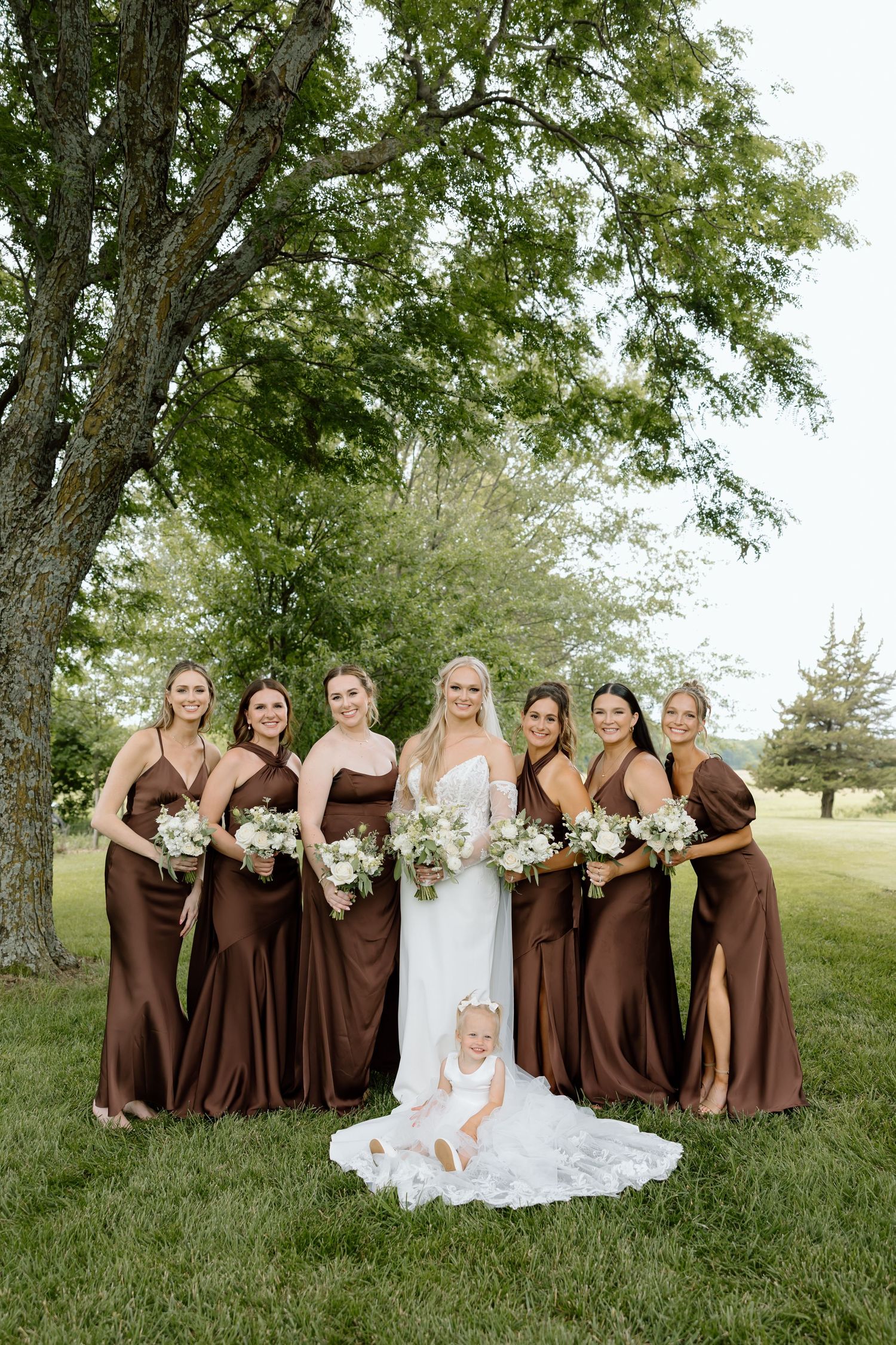 Bride in white gown with six bridesmaids in brown satin dresses holding white bouquets, posing outdoors under a large tree.