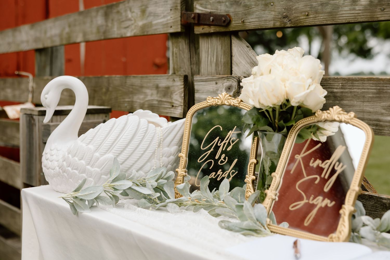 Elegant wedding gift table with white swan decor, gold framed signs, and white peony flowers near a rustic red barn.