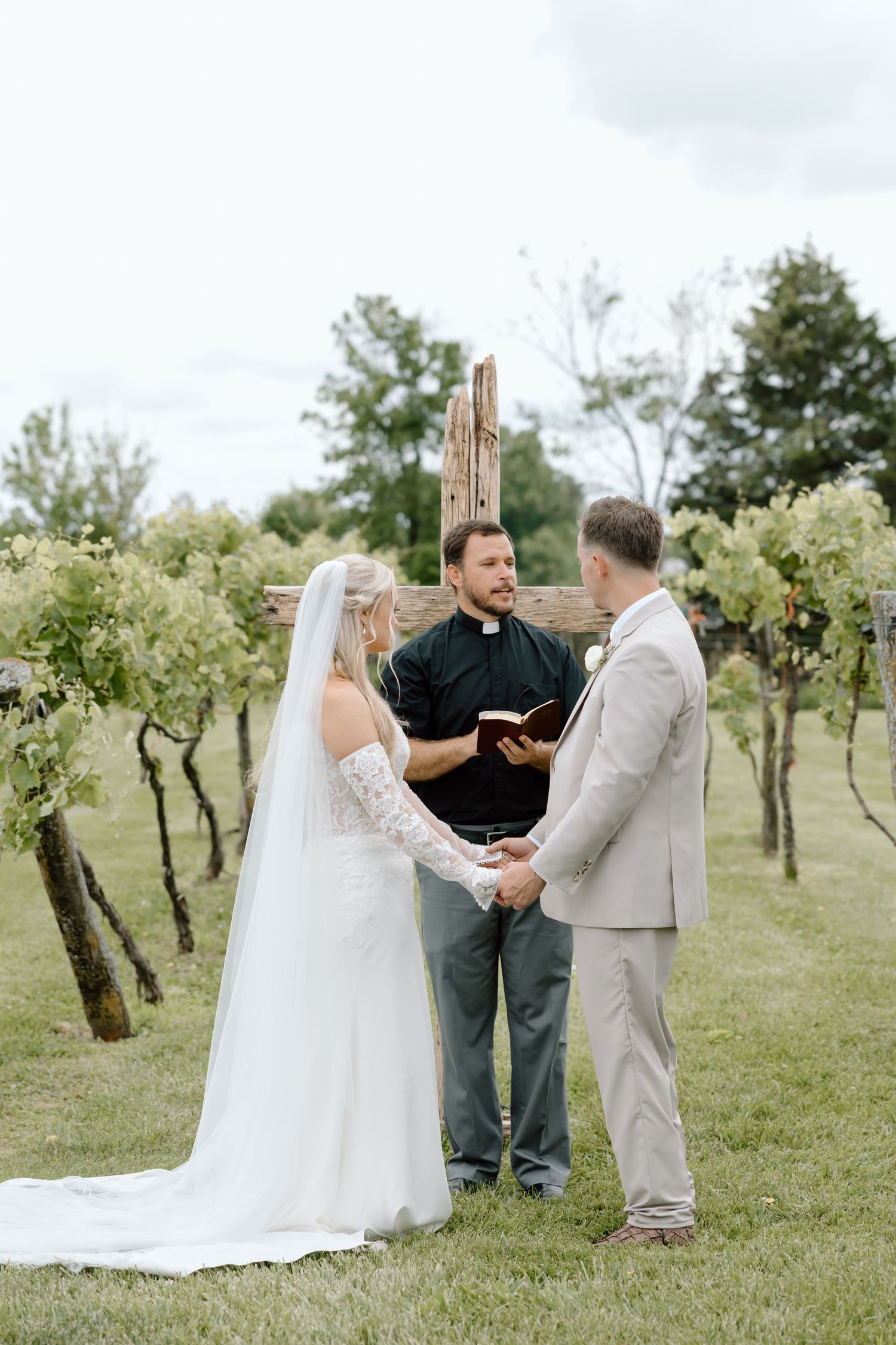 Bride and groom hold hands during outdoor vineyard wedding ceremony with officiant holding Bible.