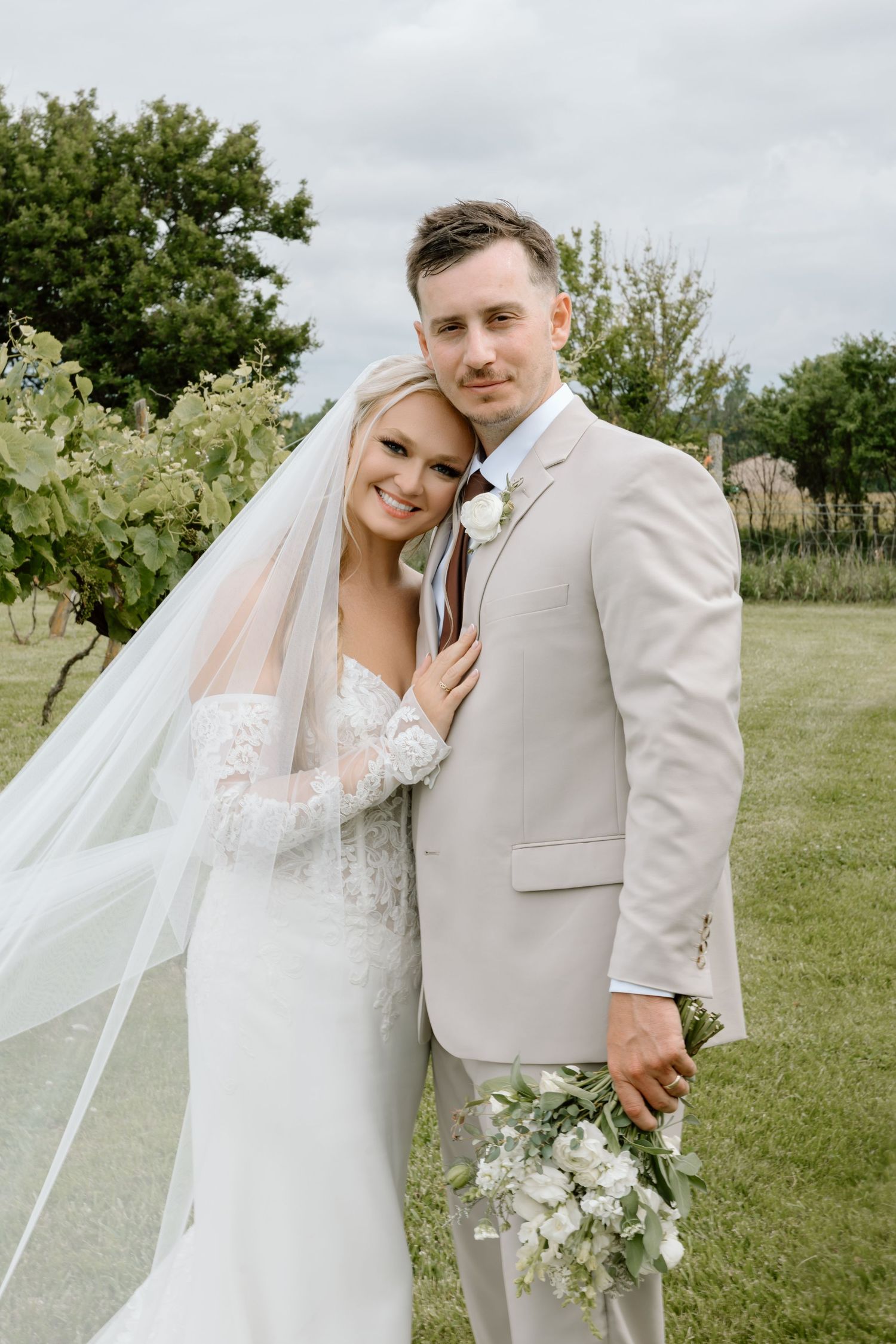Bride in lace gown and veil poses with groom in tan suit holding white bouquet outdoors at vineyard wedding.