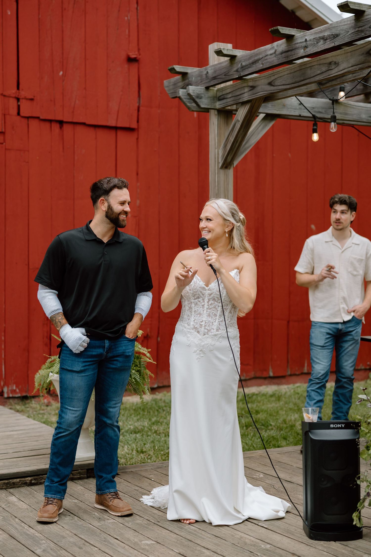 Bride in white gown speaks into microphone beside groom in black shirt at outdoor barn wedding reception.